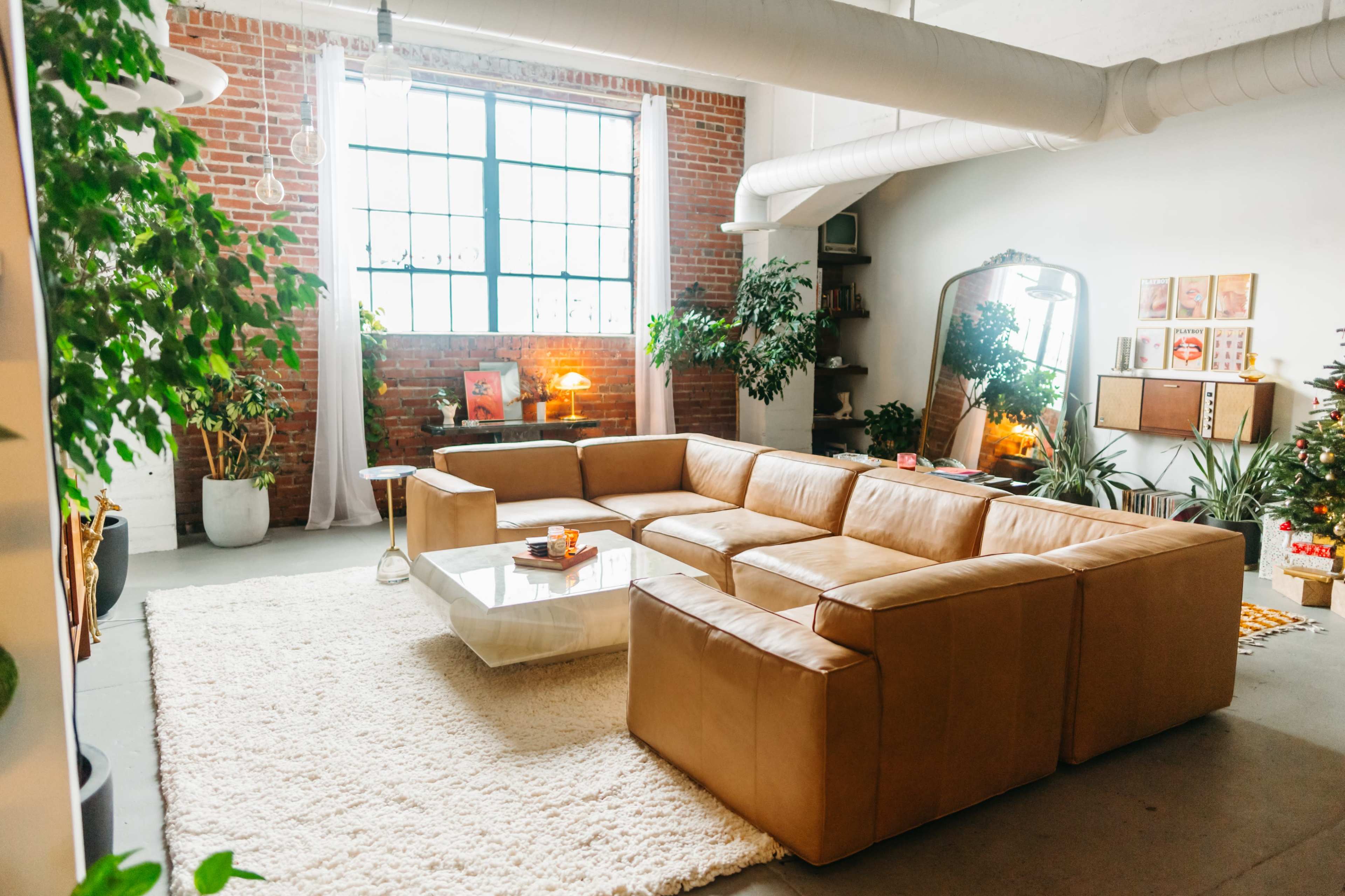 A spacious living room features a large leather sectional sofa, a glass coffee table, and plants, with a large window allowing natural light to fill the space.