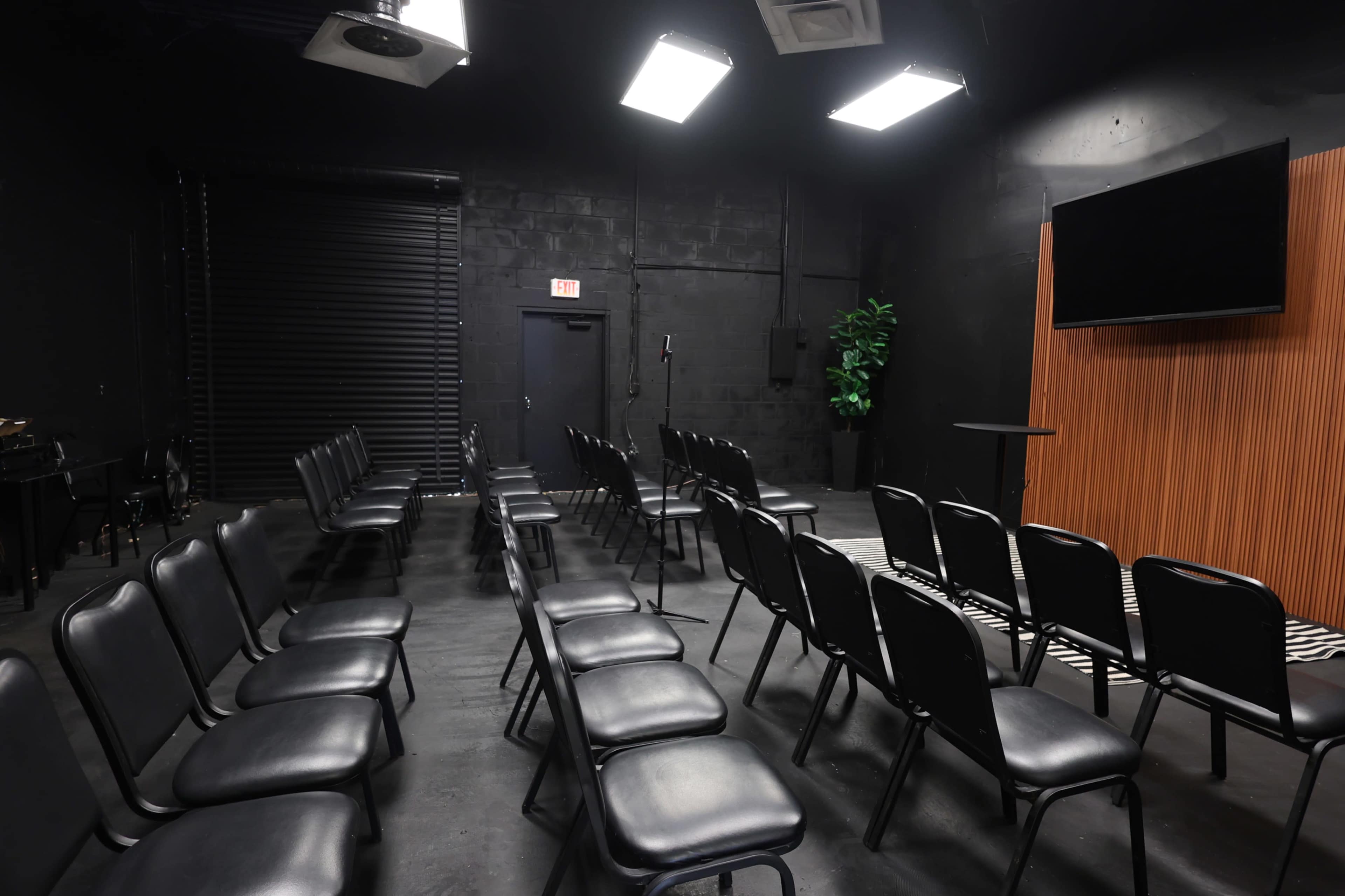 The image shows a dimly lit room set up with rows of black chairs facing a wooden panel and a monitor.