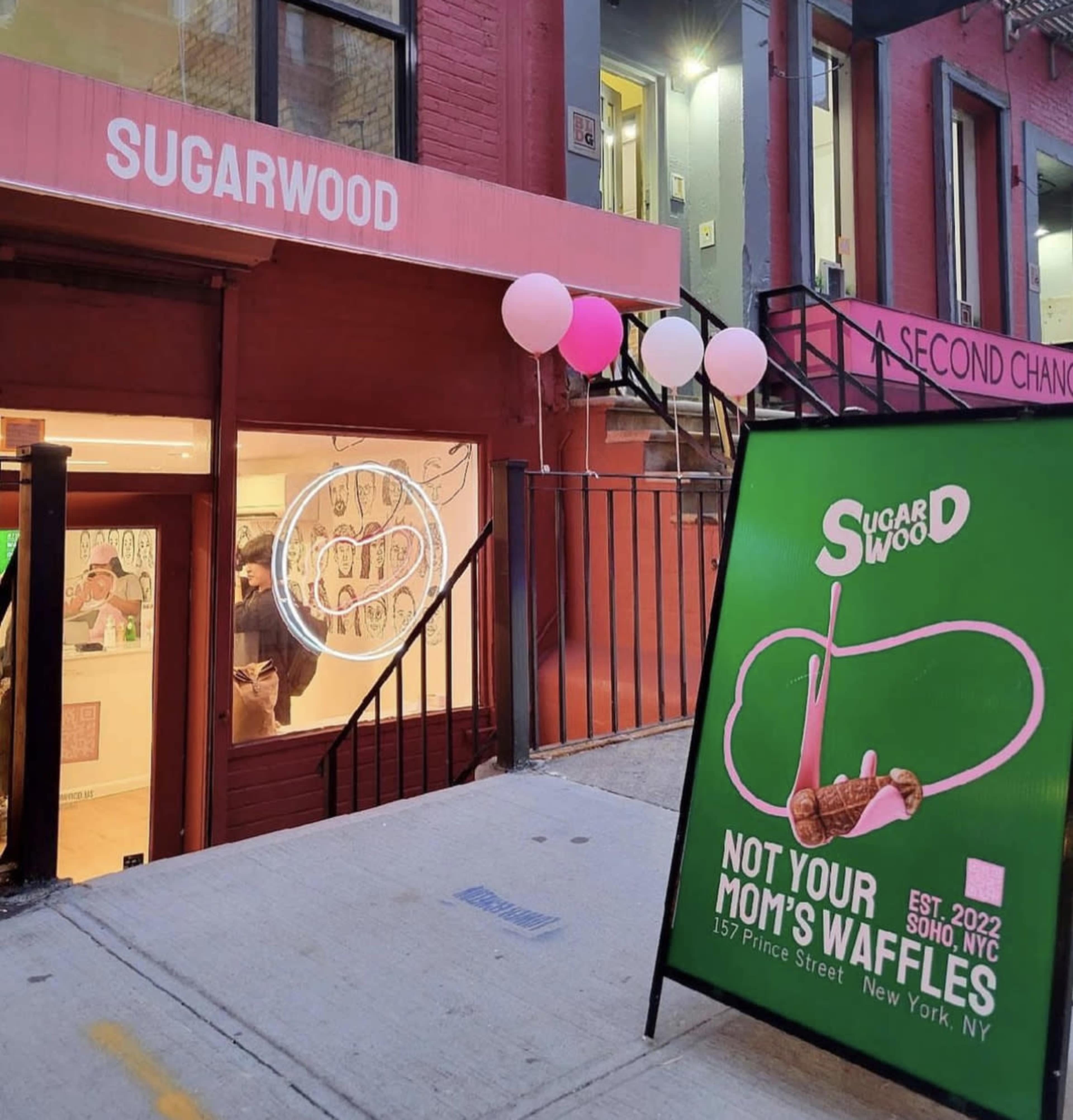 The image shows the entrance of a café named "Sugarwood," featuring a large sign advertising "Not Your Mom's Waffles" and balloons decorating the exterior on a city street in New York.