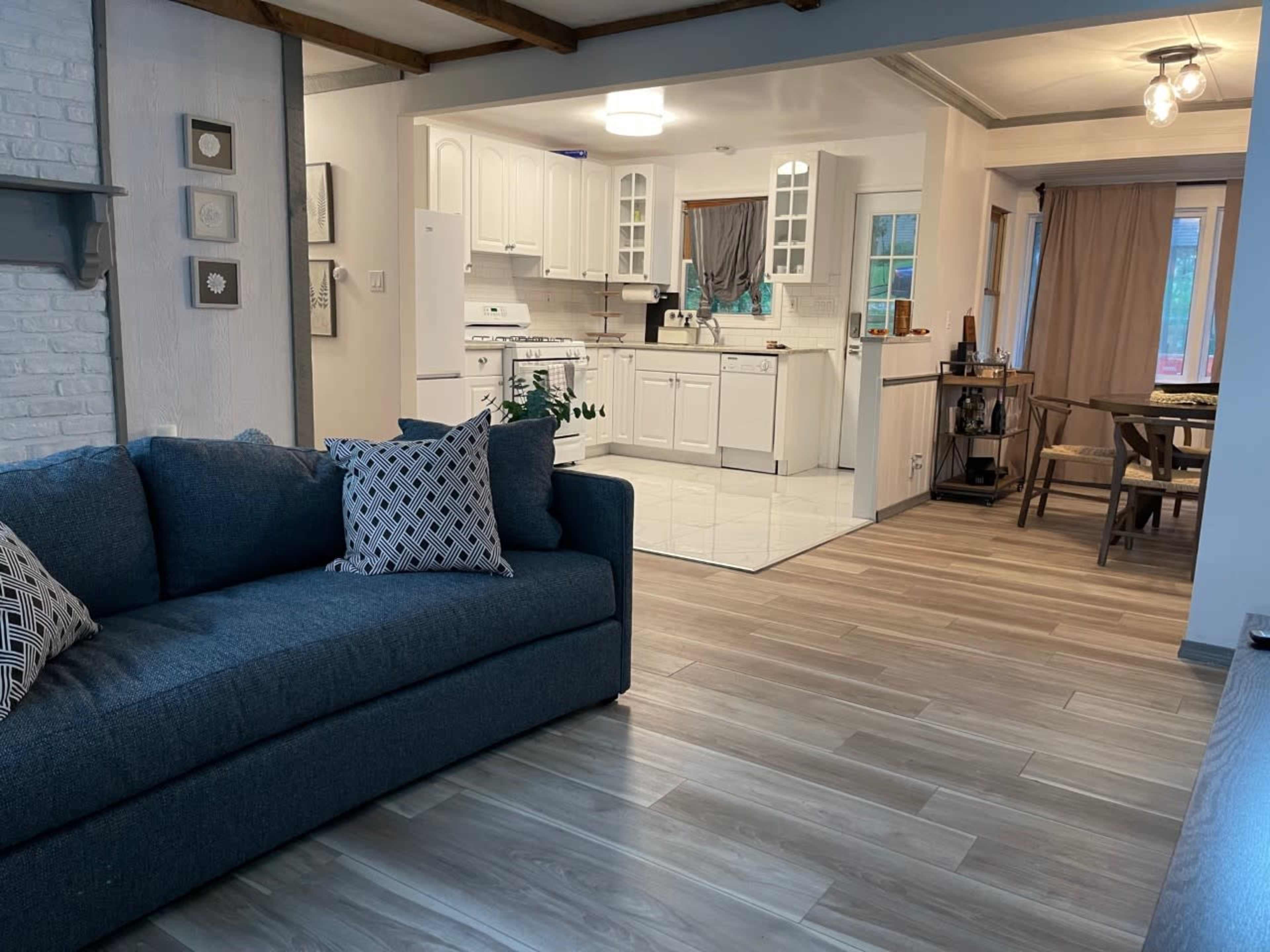 A cozy living area with a dark blue sofa and a view of a spacious kitchen and dining area featuring white cabinetry and a wooden dining table.
