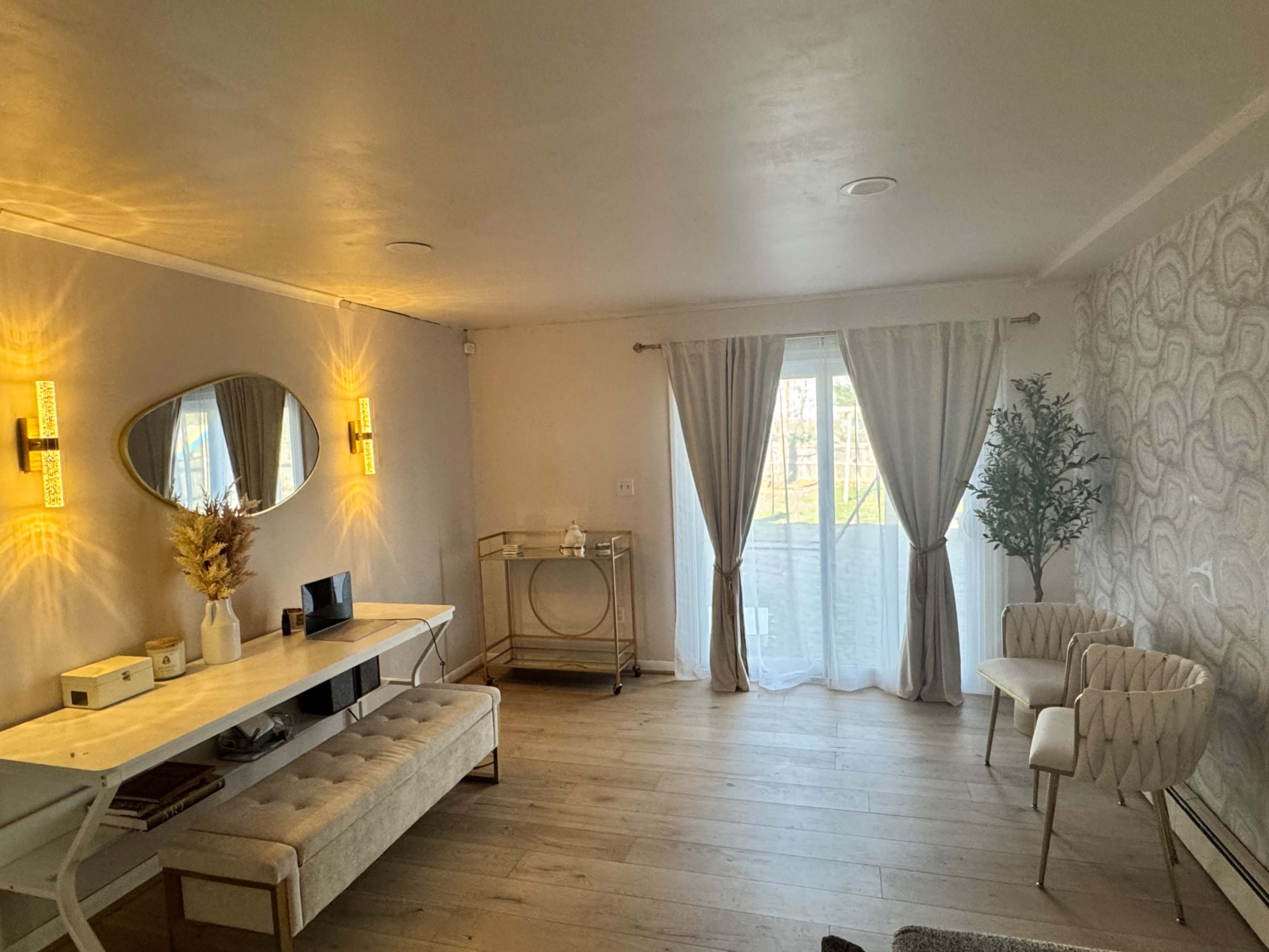The image shows a modern, minimalist living room with a dining table, a decorative mirror, and large windows covered by sheer curtains, allowing natural light to fill the space.