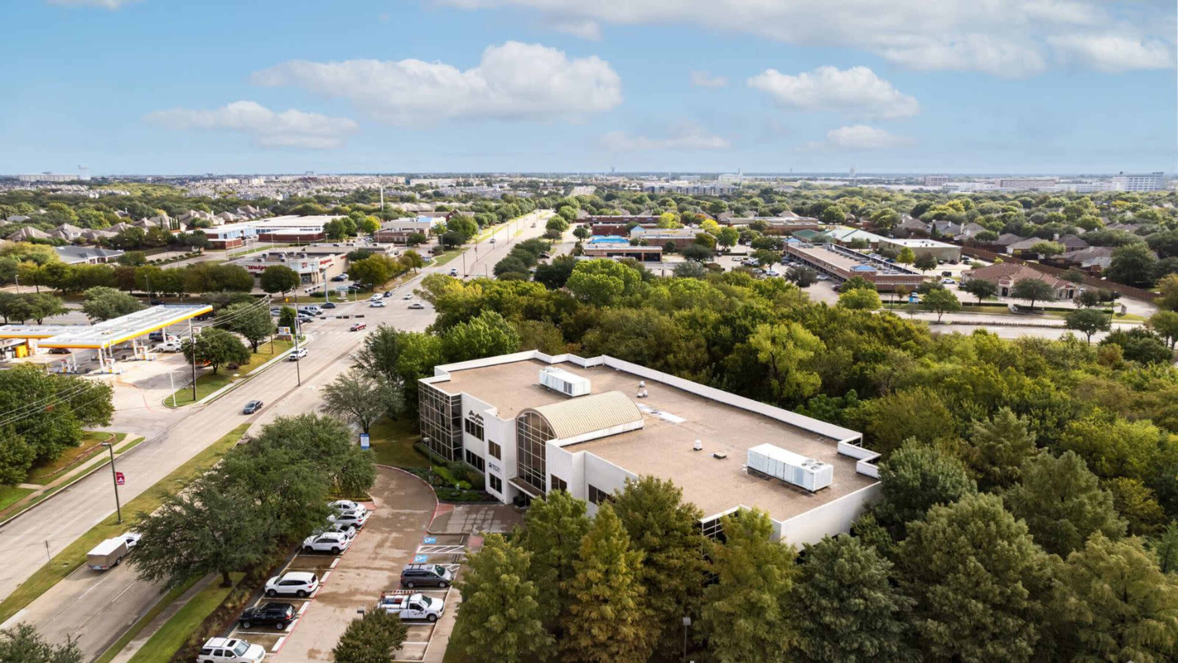 A view of a commercial building surrounded by trees and residential areas, with a road and vehicles visible nearby.