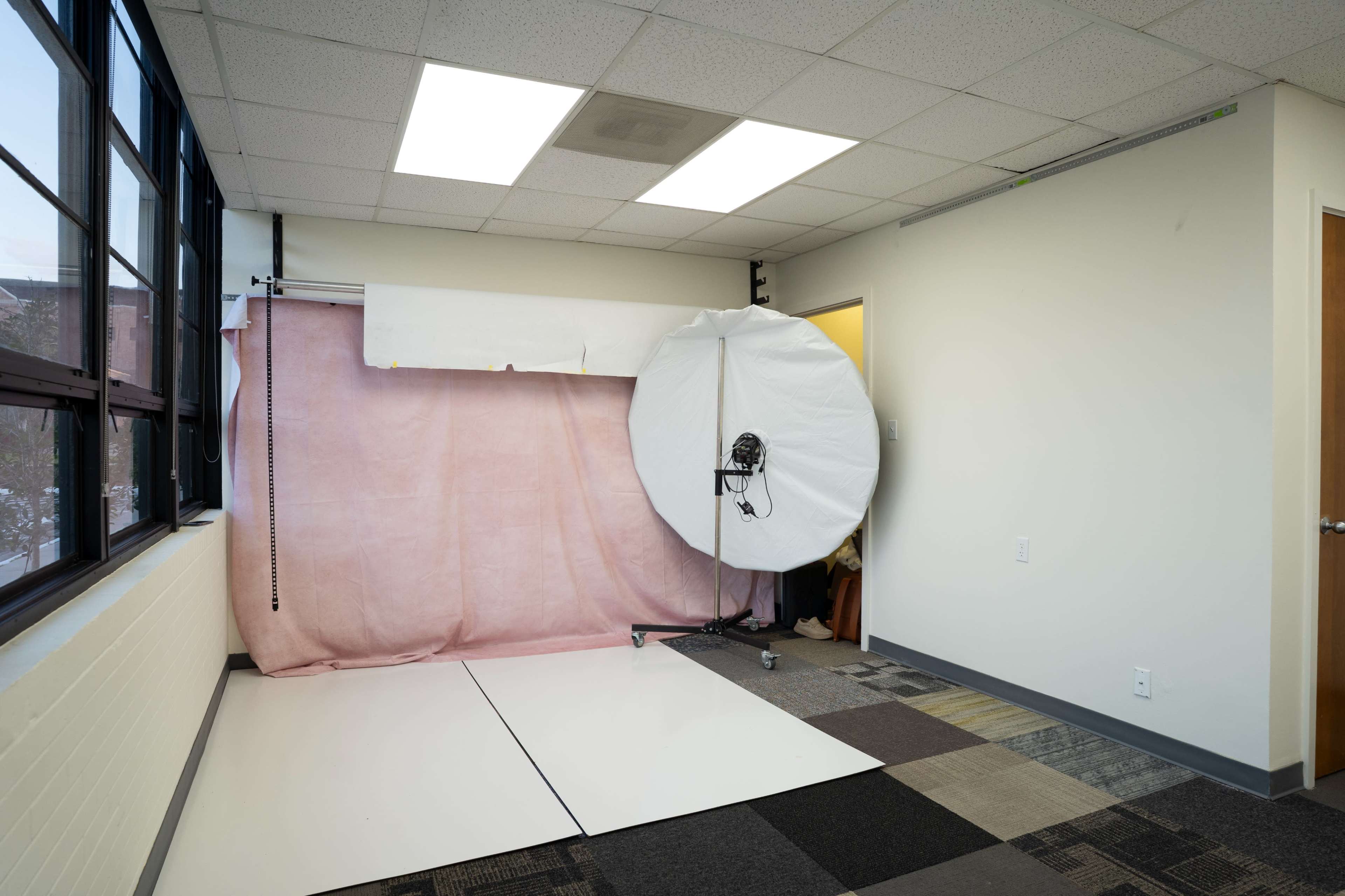 The image shows an empty room equipped with a light setup, a pink backdrop, and a section of carpeted flooring.