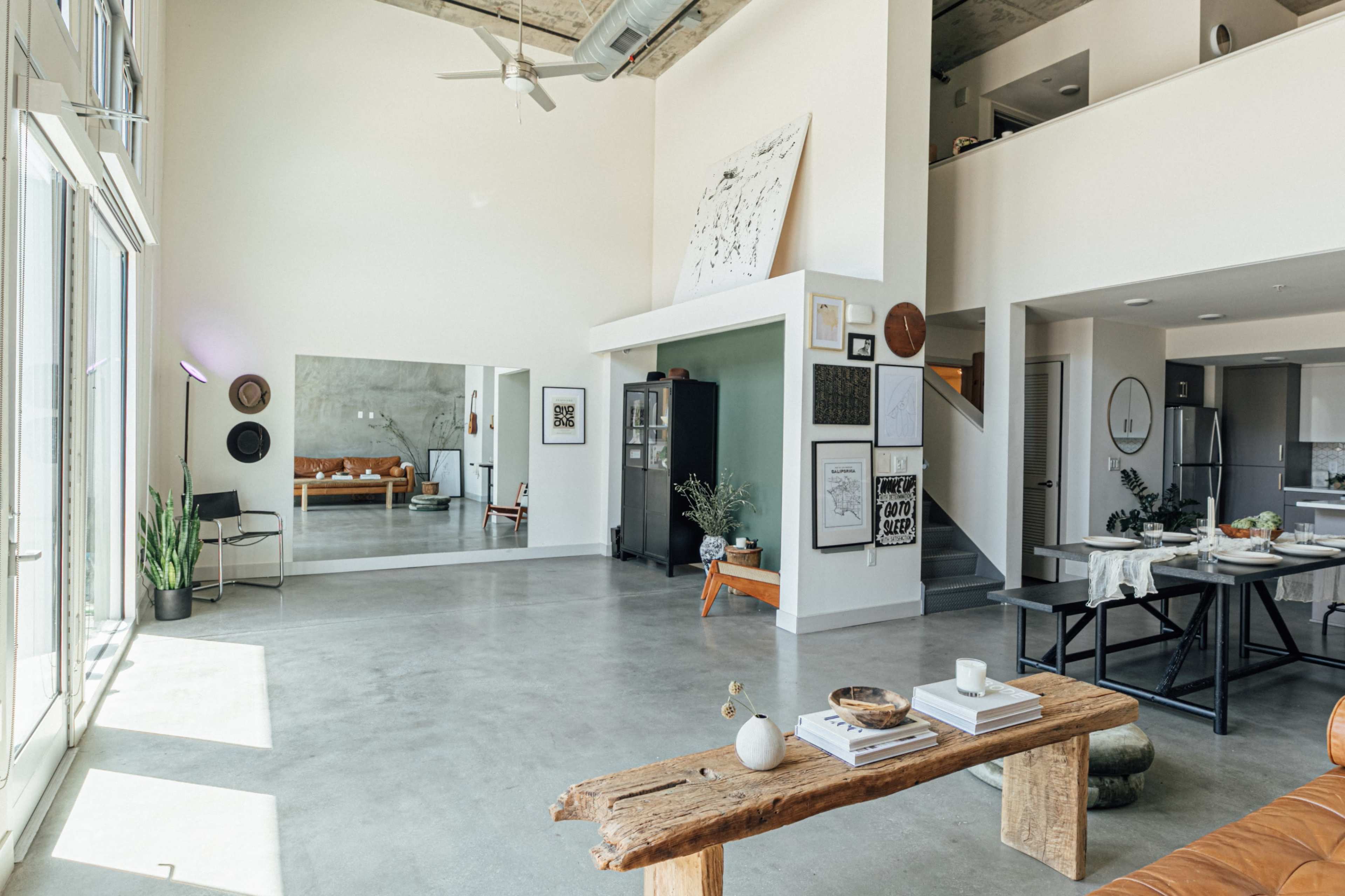 A spacious open-concept living area features a large mirror, a wooden table, and various wall art, illuminated by natural light from the large windows.
