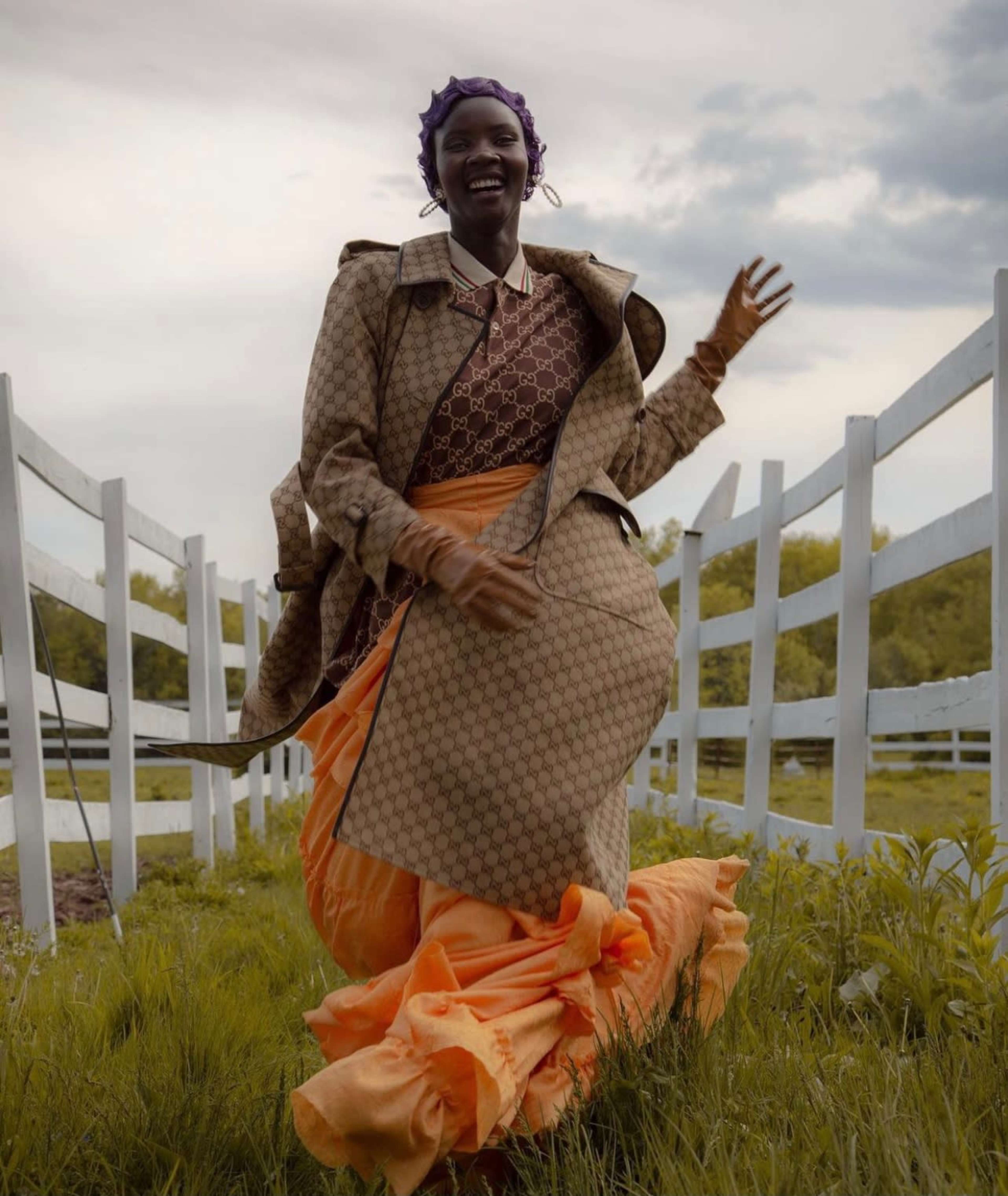 A woman wearing a patterned coat and a flowing orange skirt walks joyfully along a grassy path beside a white fence.