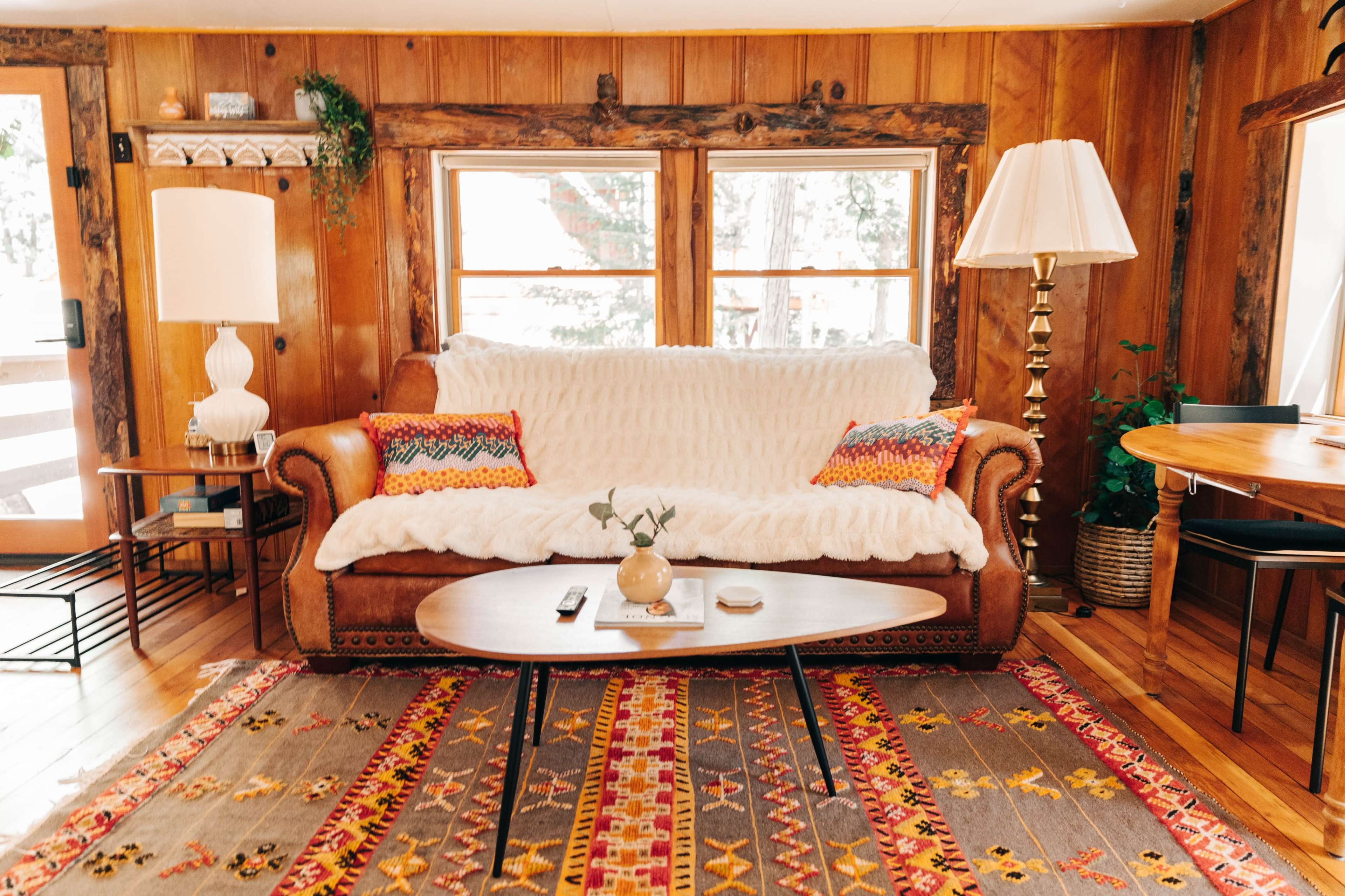 The image shows a cozy living room with a brown leather sofa covered by a white throw, decorative pillows, a coffee table, and a patterned rug on the wooden floor.