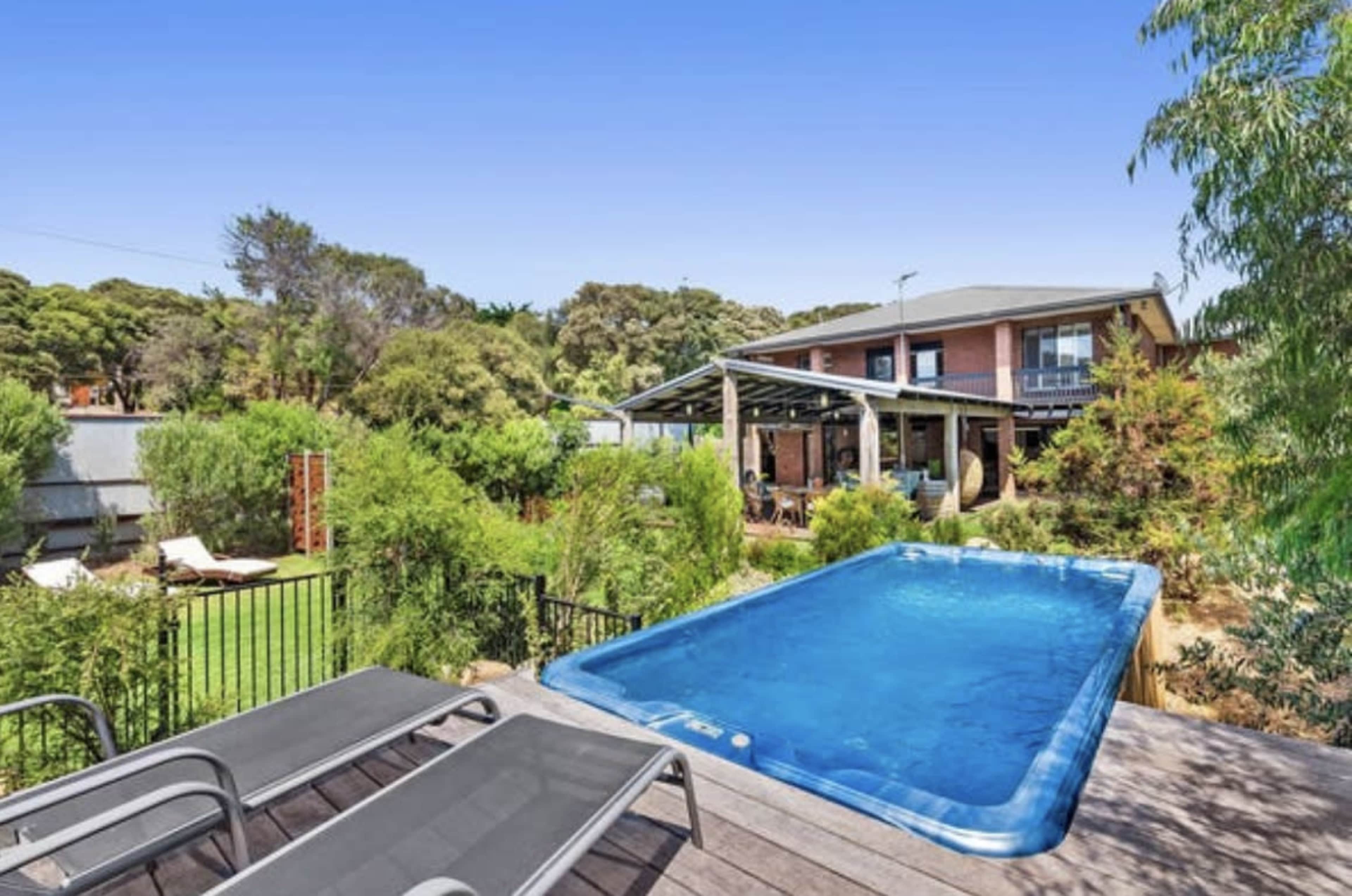 A blue swimming pool is situated in a backyard, surrounded by greenery and overlooking a two-story brick house.