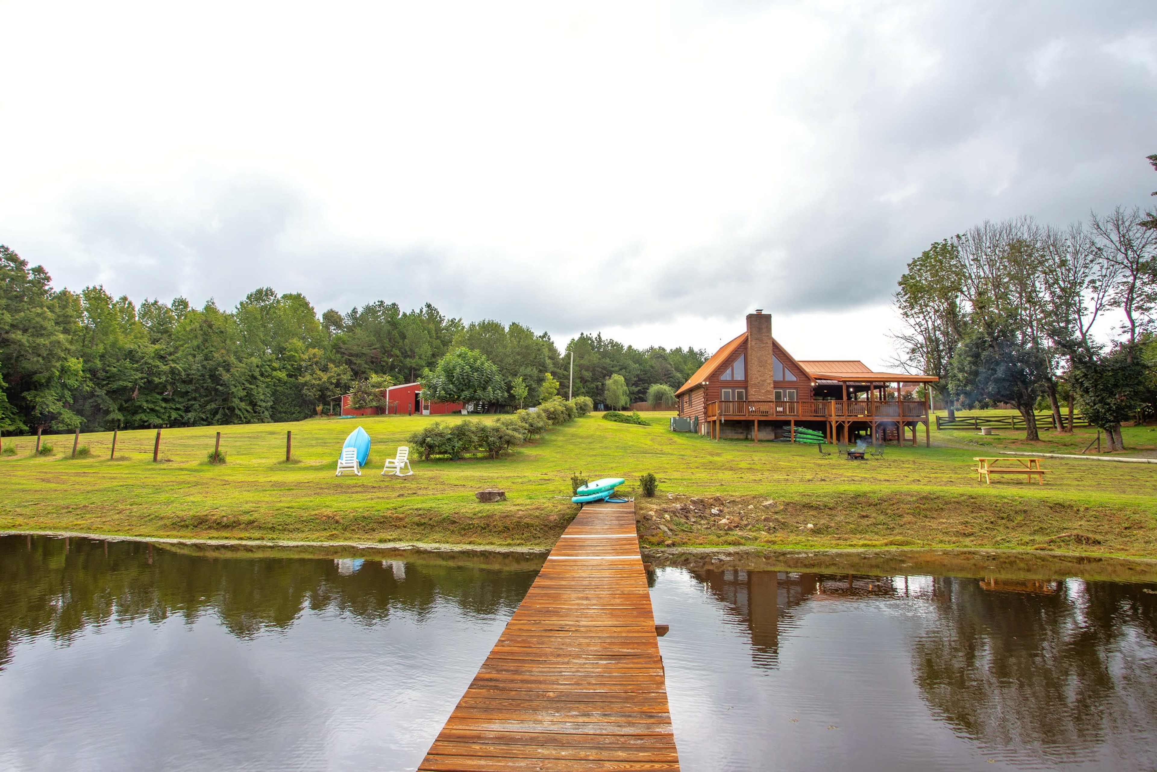 A wooden dock extends over a calm body of water, leading to a two-story house set against a backdrop of greenery under a cloudy sky.
