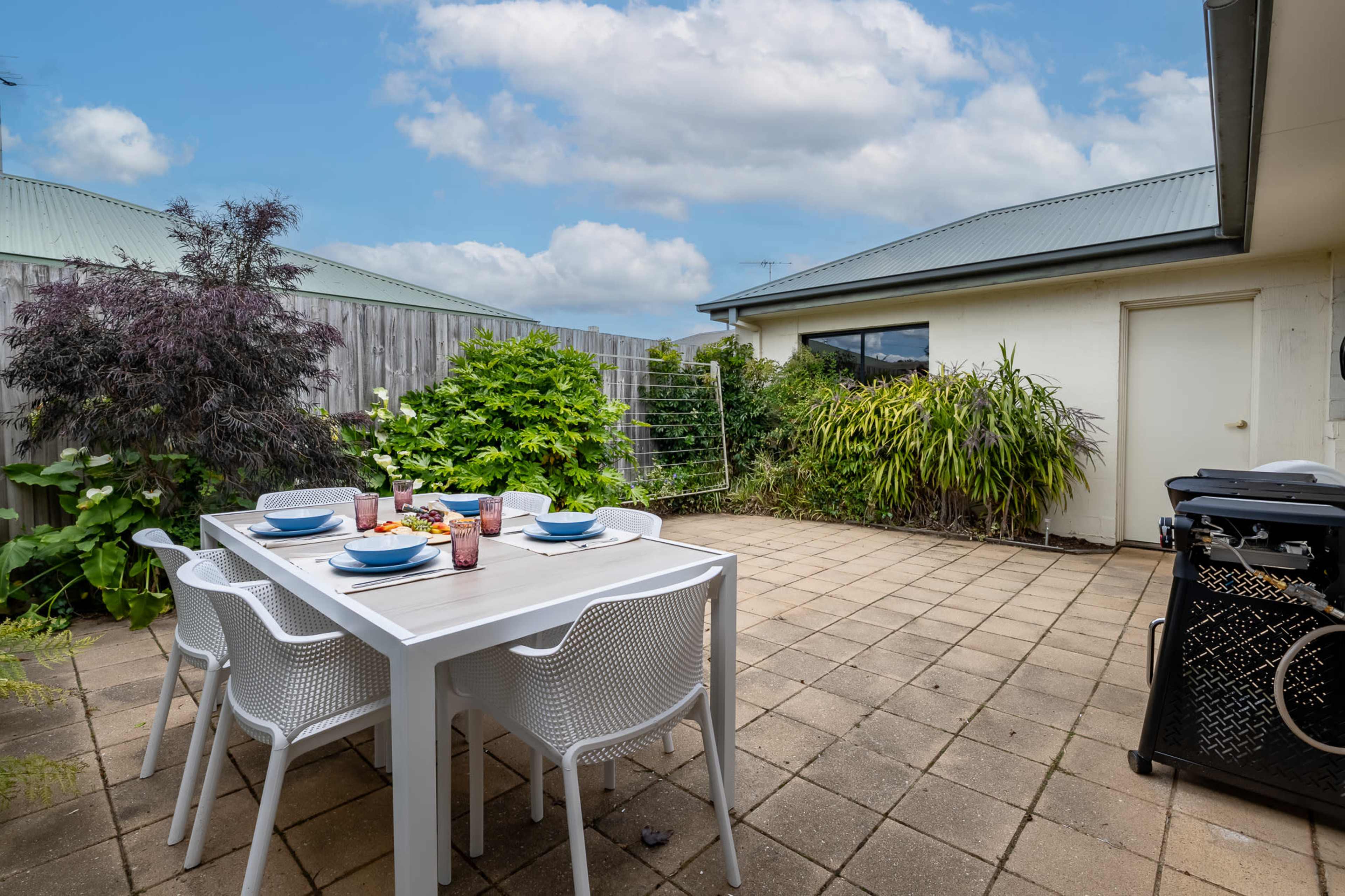 The image shows a patio area featuring a table set for a meal with plates and drinks, surrounded by greenery and a house in the background.