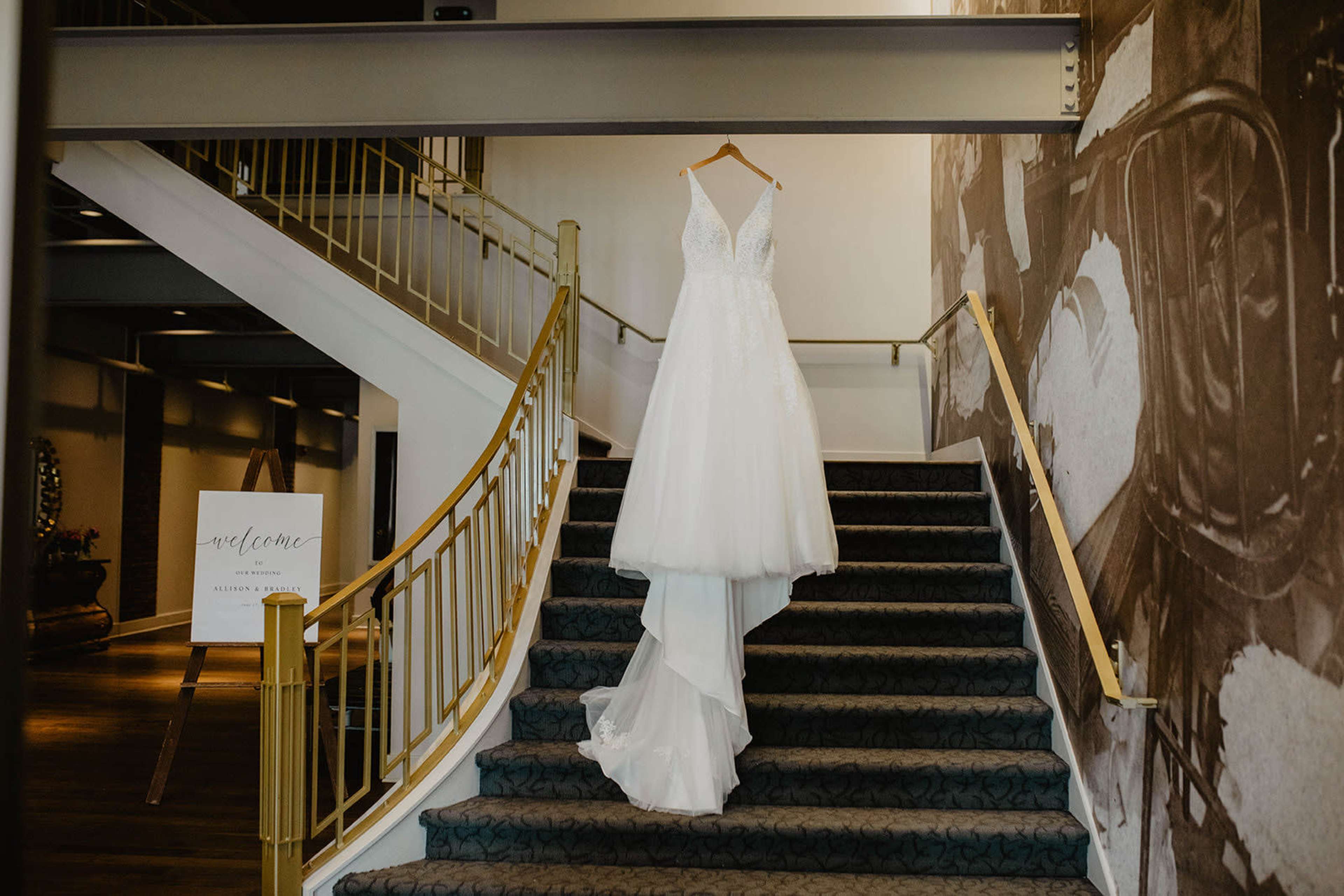 A wedding dress is displayed on a hanger at the top of a staircase in a venue with a welcoming sign nearby.