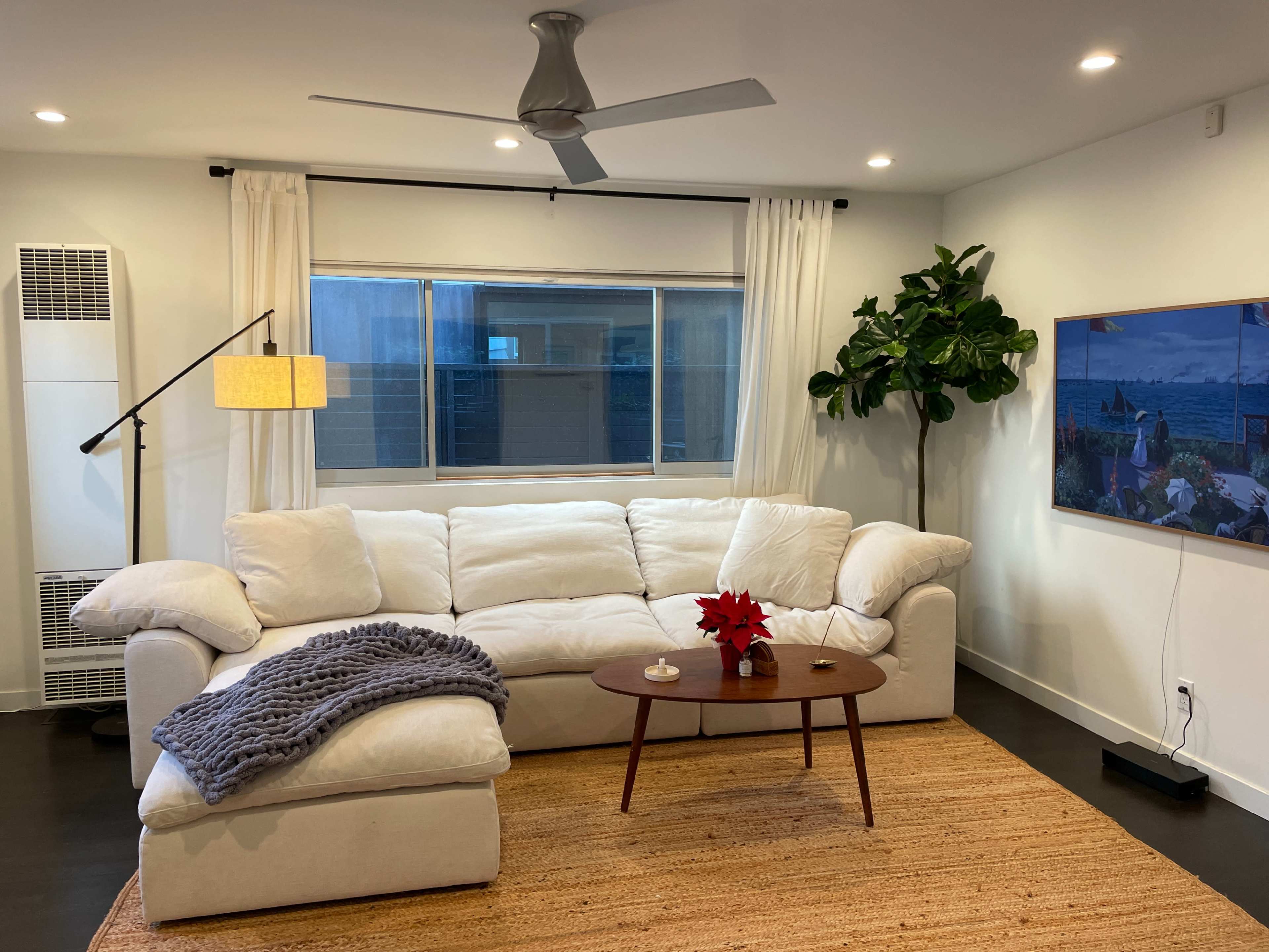 The image shows a modern living room featuring a white sectional sofa with a throw blanket, a round coffee table, a large window, and a green indoor plant.