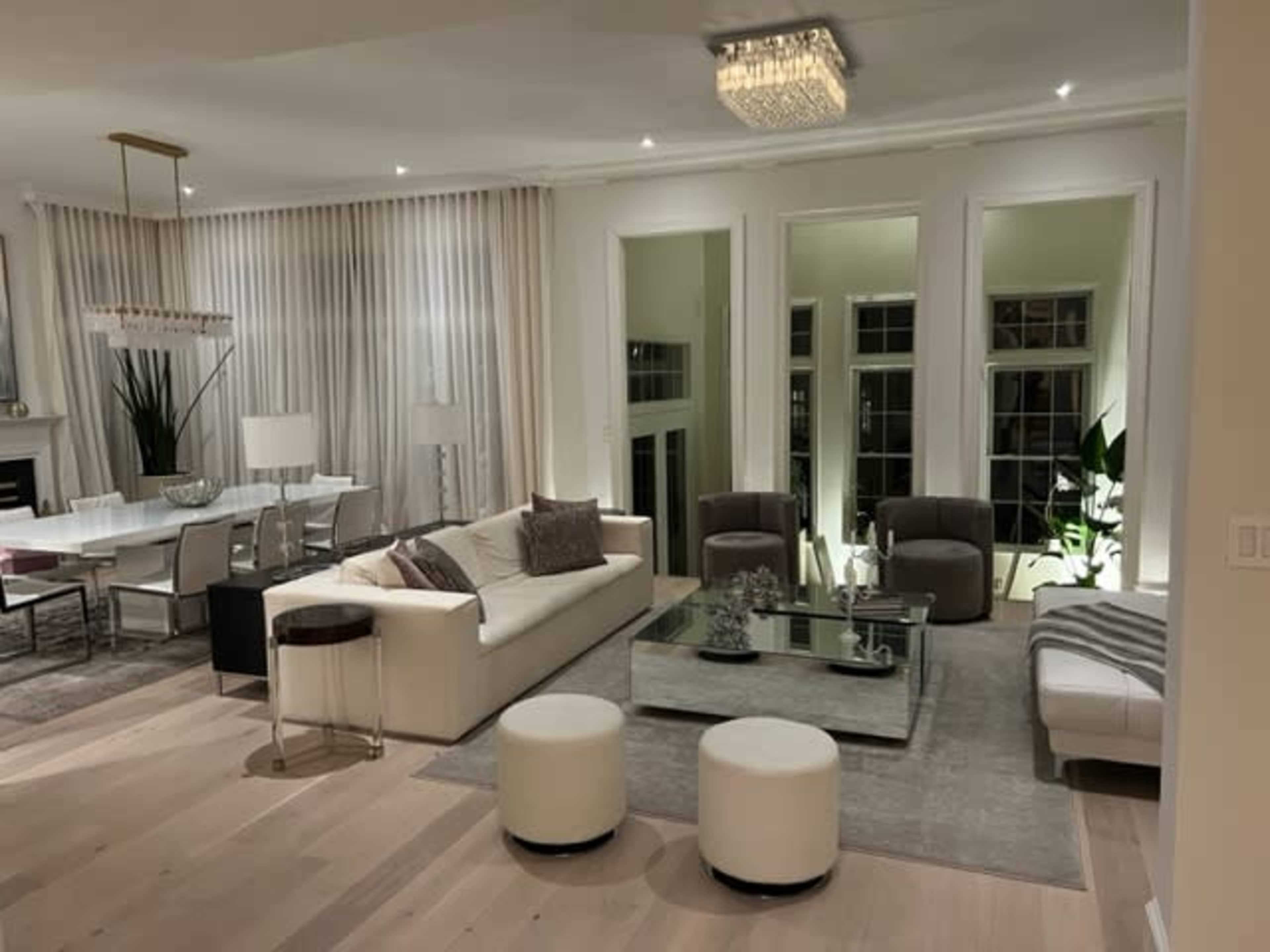 A stylish living room features a white sofa, glass coffee table, two armchairs, large windows, and a chandelier, with modern decor accents.