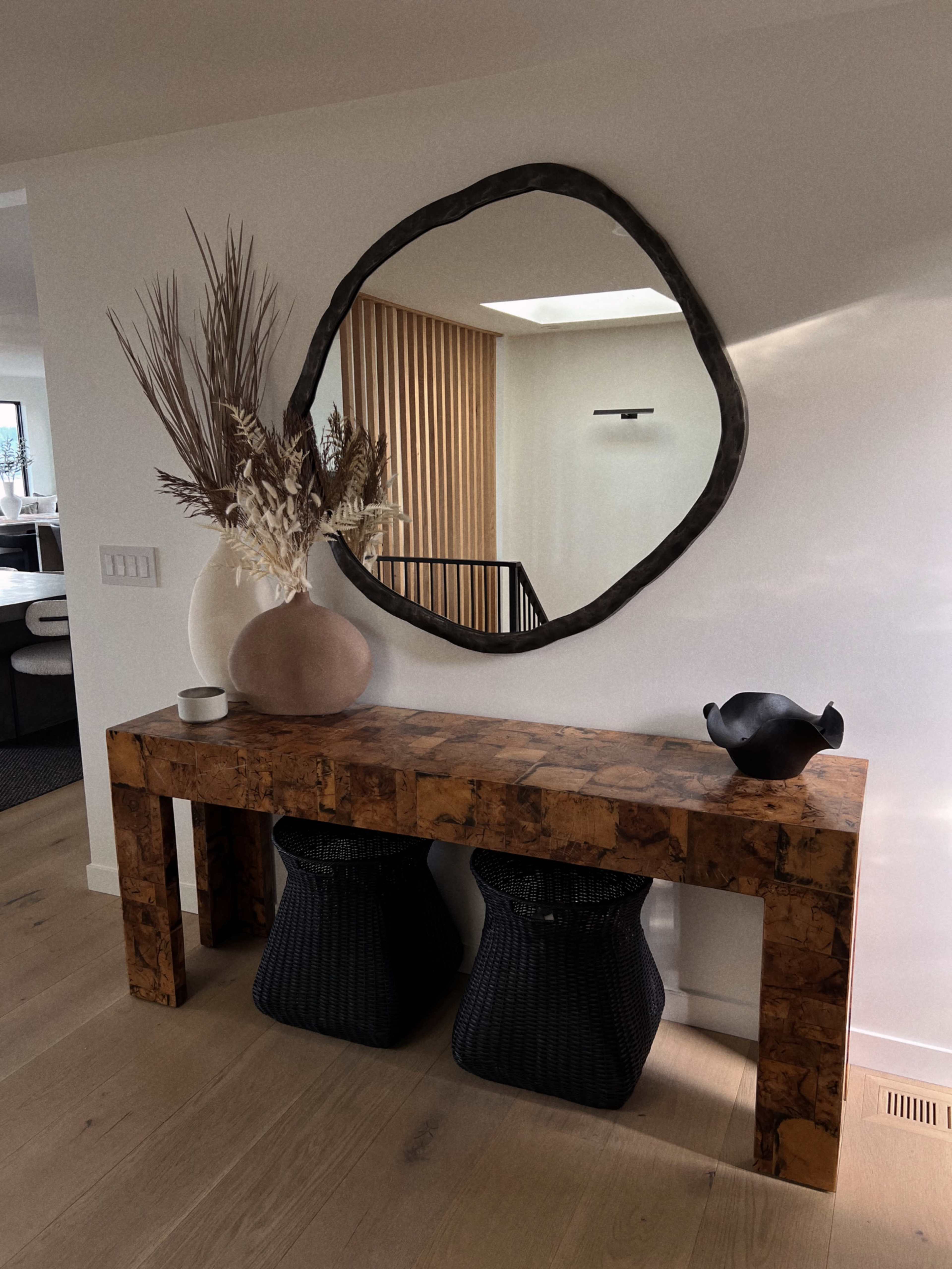 A wooden console table holds a large vase with dried plants and a small bowl, accompanied by two black woven baskets underneath and a round mirror above.