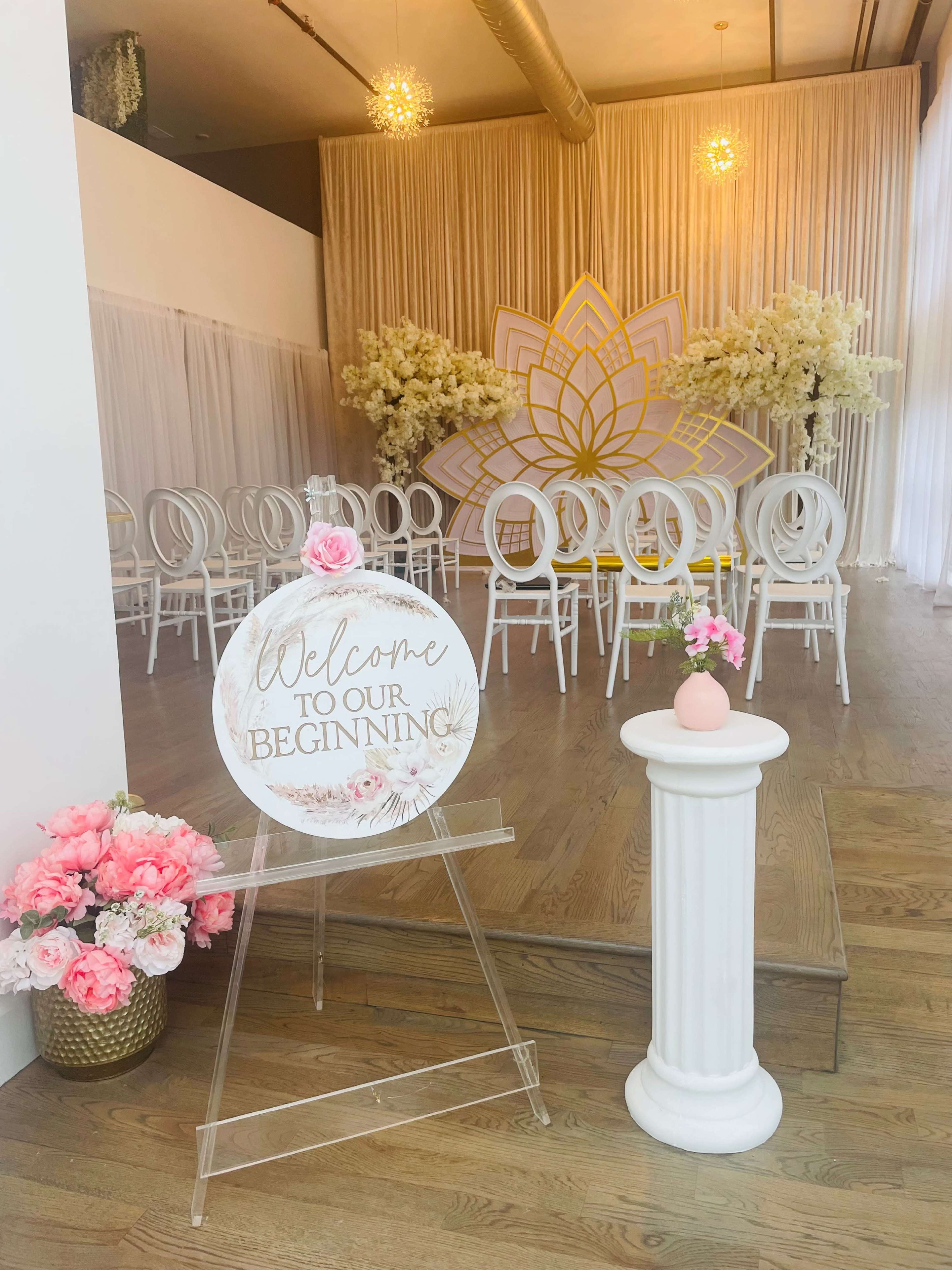 A welcome sign adorned with flowers stands next to white chairs arranged in front of a floral backdrop in a decorated event space.