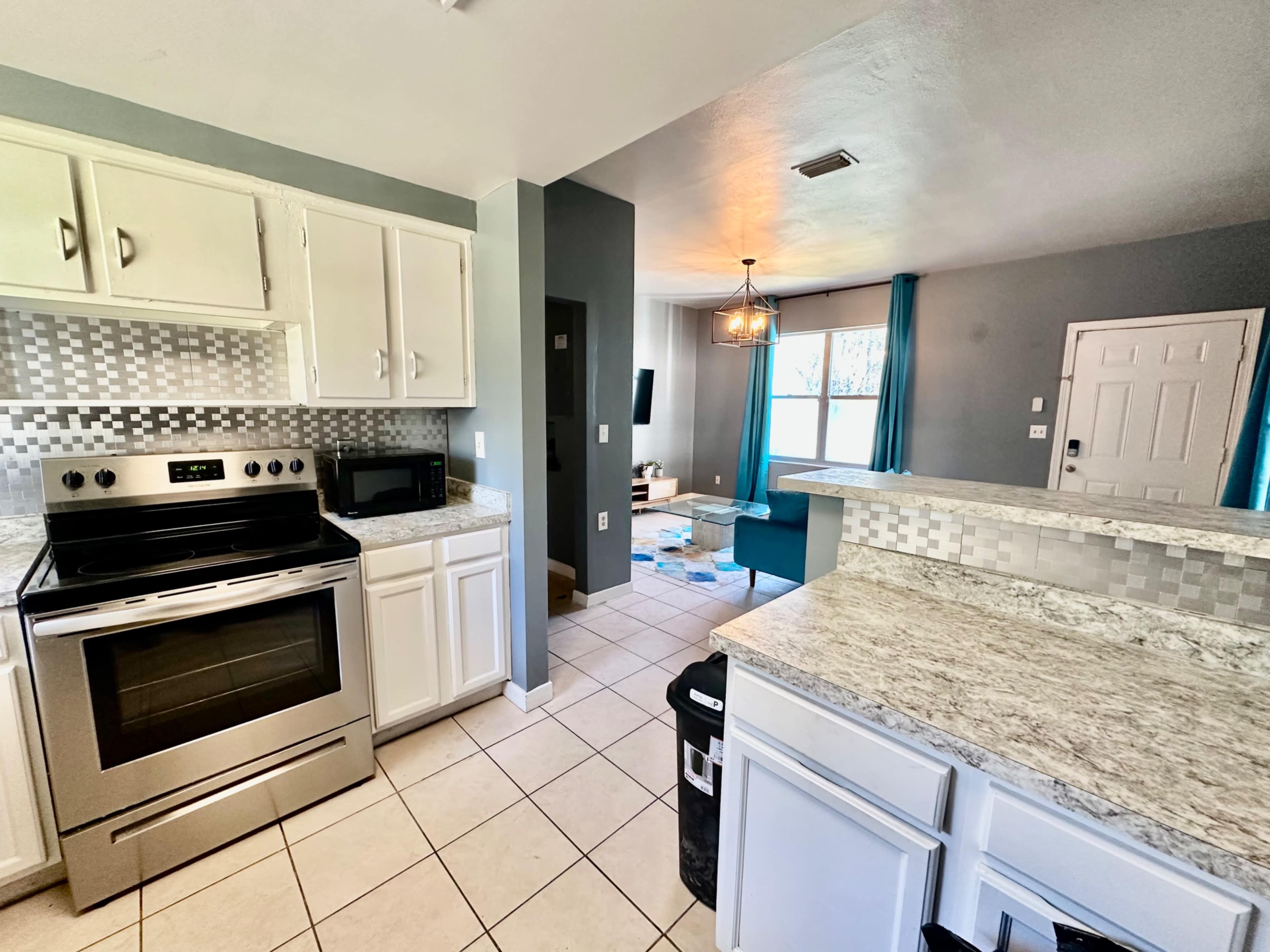 The image shows a modern kitchen with white cabinets, granite countertops, and stainless steel appliances, leading into a living area with blue curtains and a television.