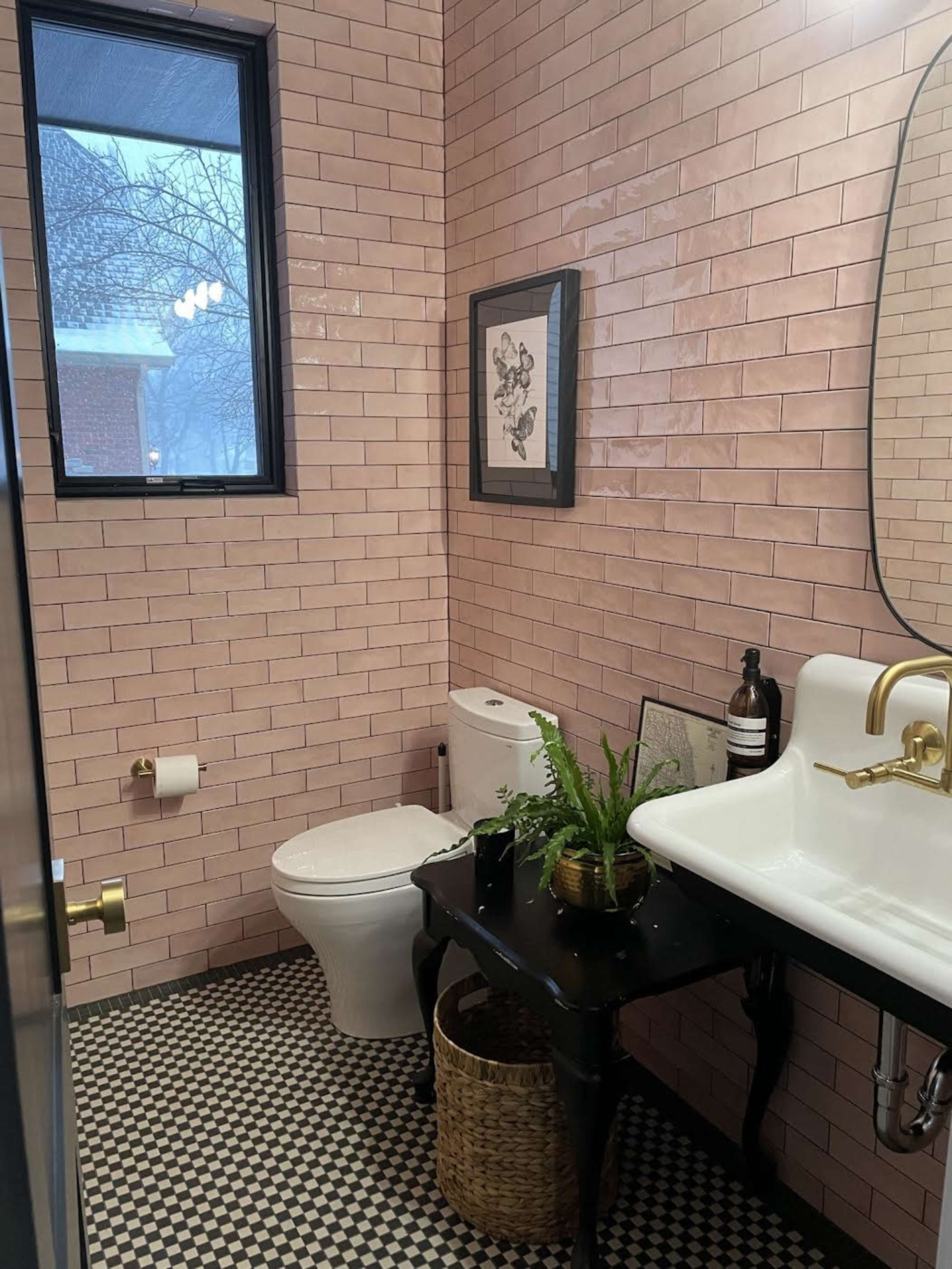 A small bathroom featuring pink tiled walls, a white toilet, a vintage sink, and a table with a plant and decorative items.
