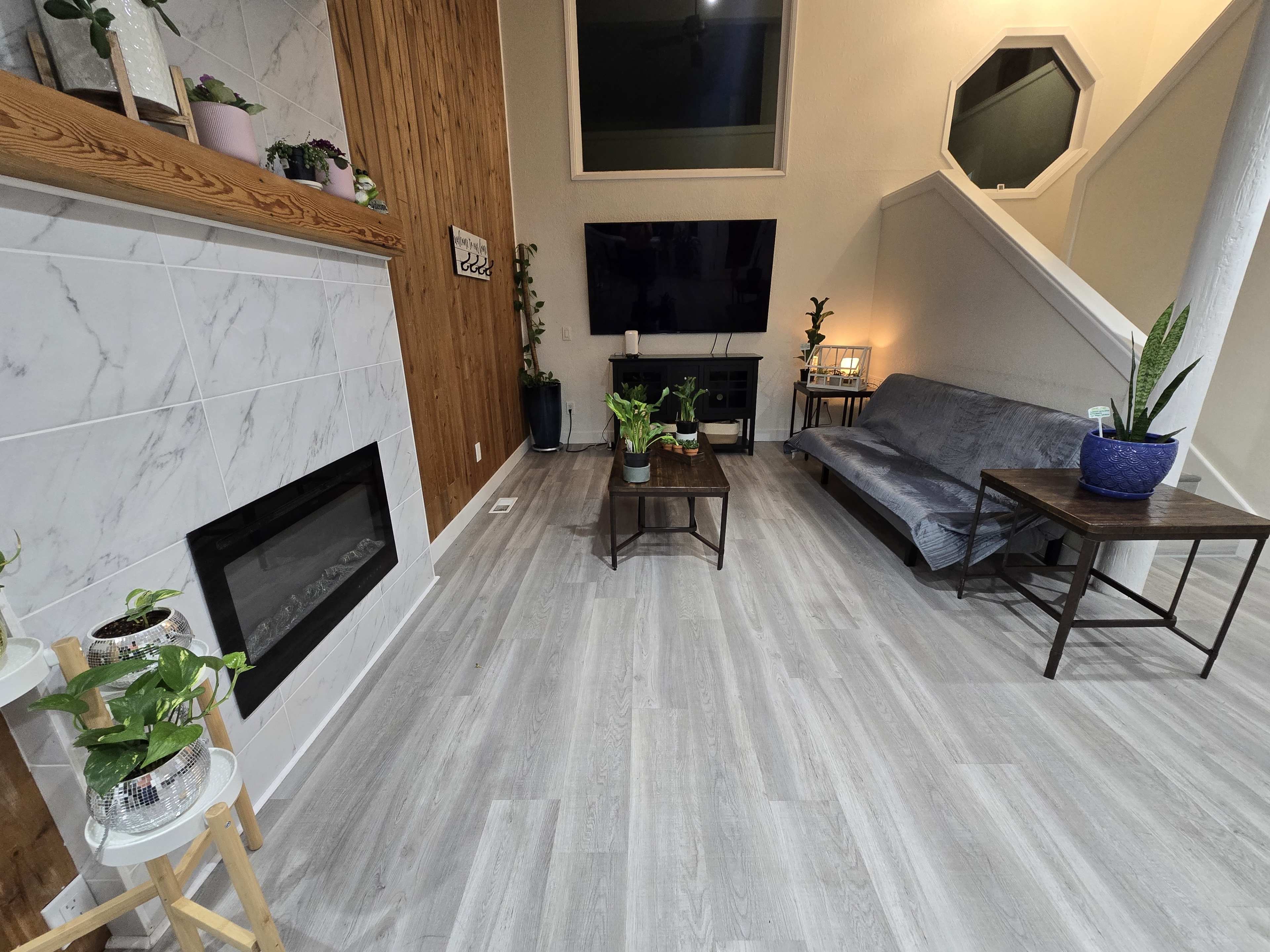 The image shows a modern living room with a wooden accent wall, a sofa, a coffee table, and various indoor plants arranged around a wall-mounted television and a faux fireplace.