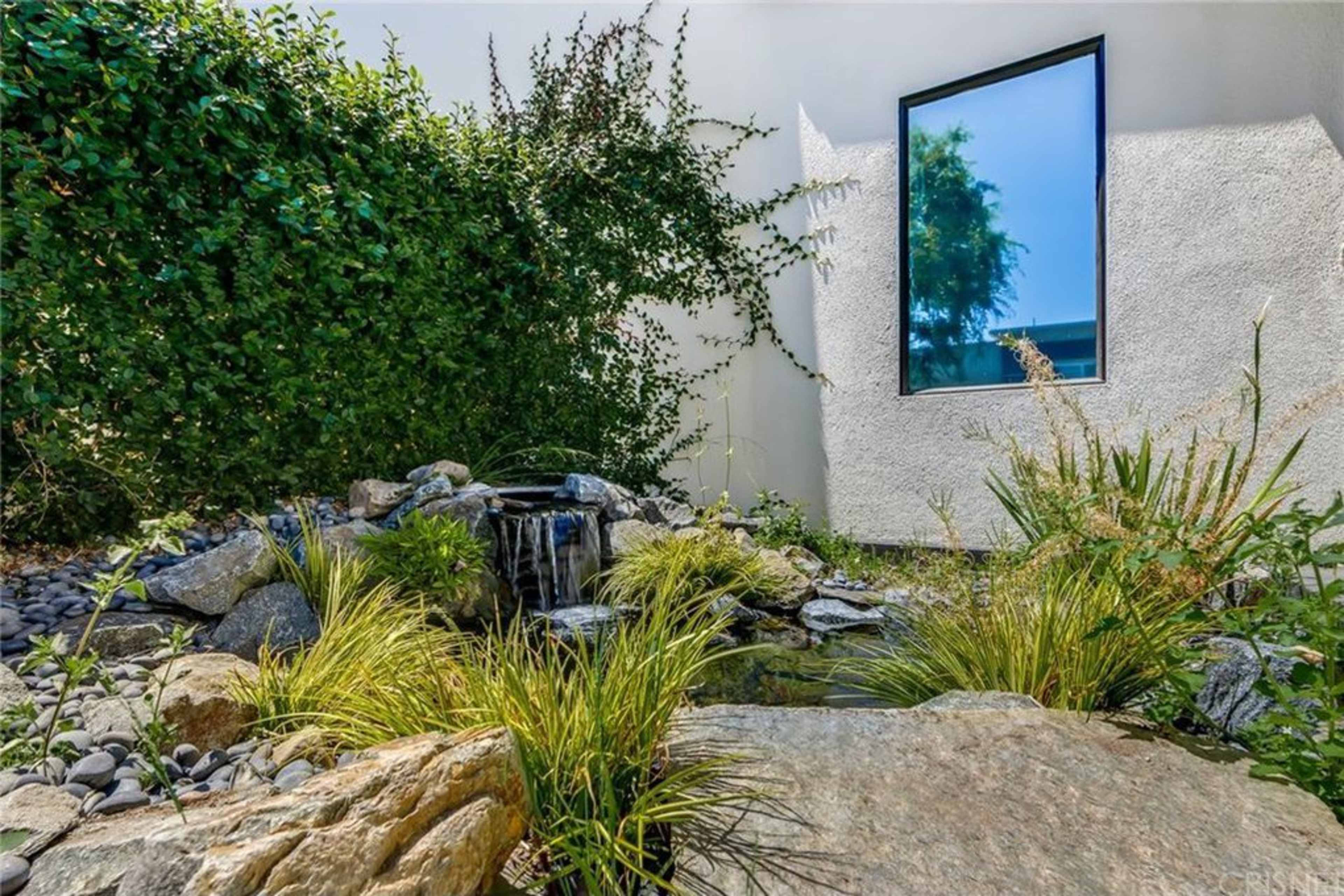 The image shows a landscaped area featuring a small waterfall flowing into a pond, surrounded by rocks and greenery, with a window visible in the background.