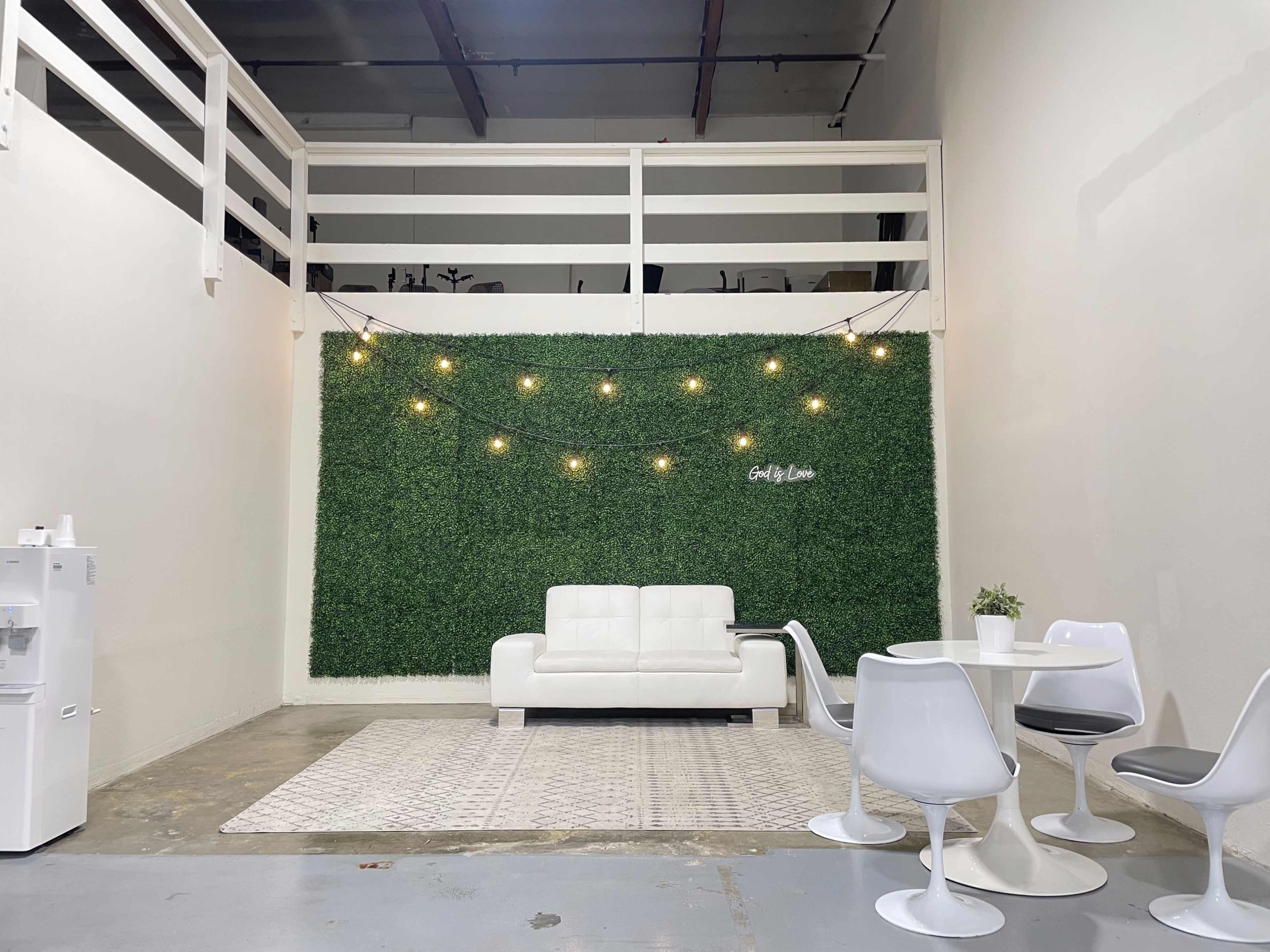 A modern interior space featuring a white sofa against a green wall of artificial grass, illuminated by string lights, with a dining table and chairs nearby.