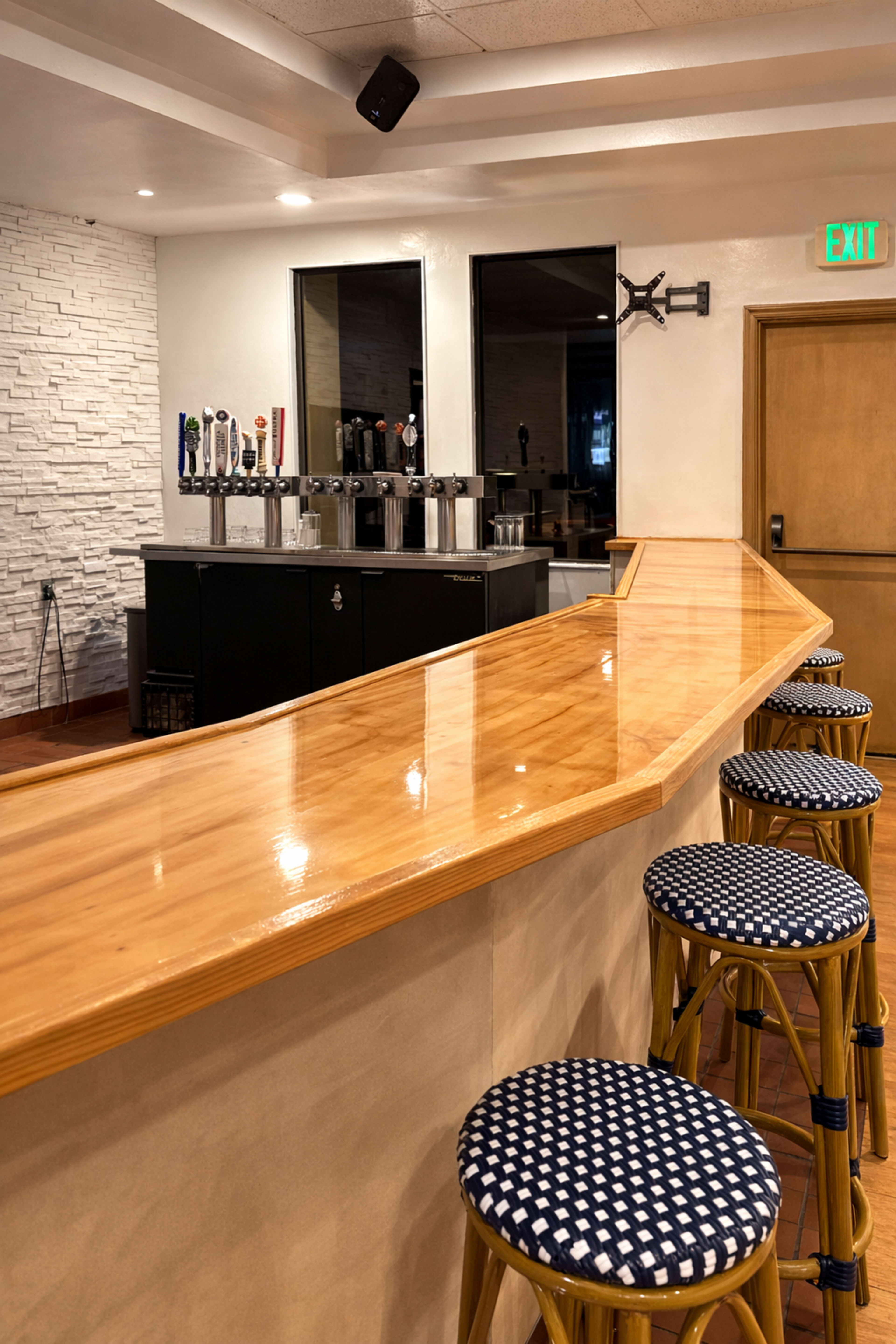 The image shows a clean, modern bar area featuring a polished wooden counter, several taps for beverages, and blue-and-white patterned stools lined up in front.