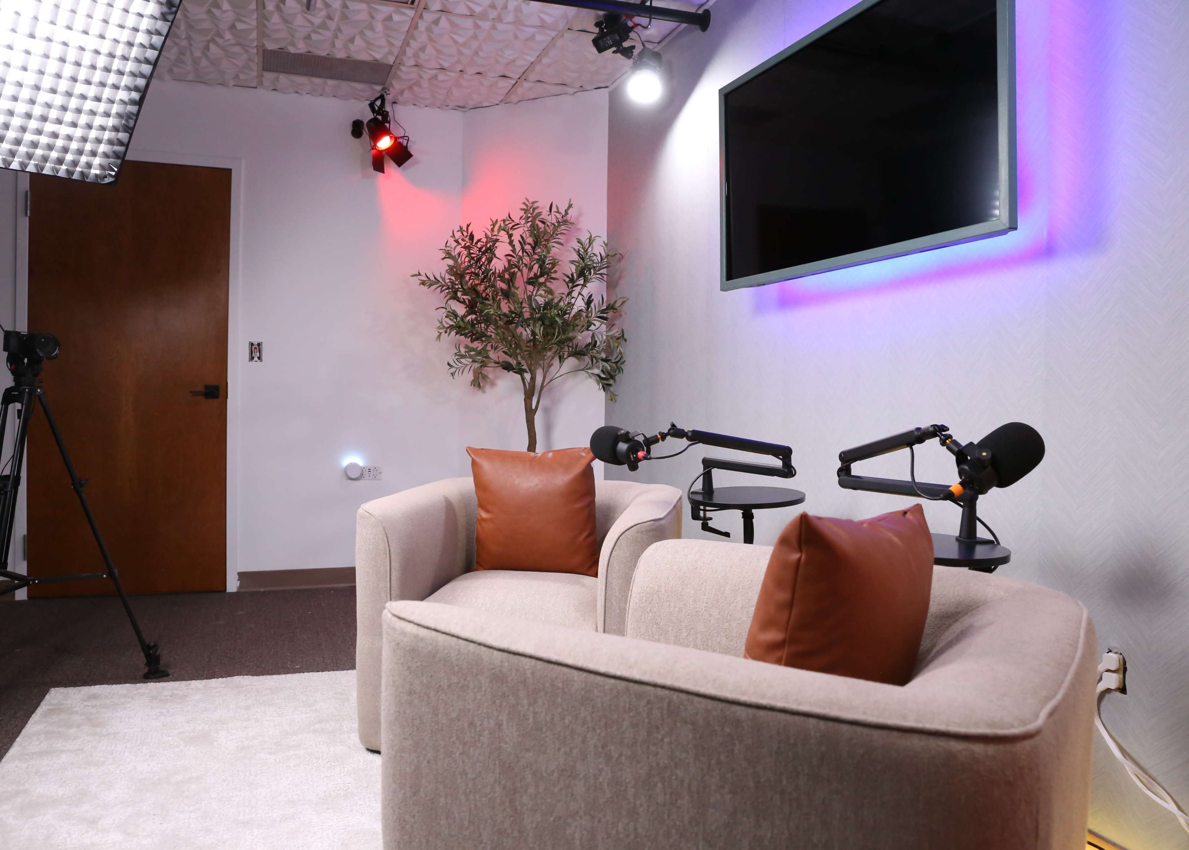 A small interview setup features two armchairs with brown cushions, a plant, and a large screen on the wall, illuminated by soft lighting.