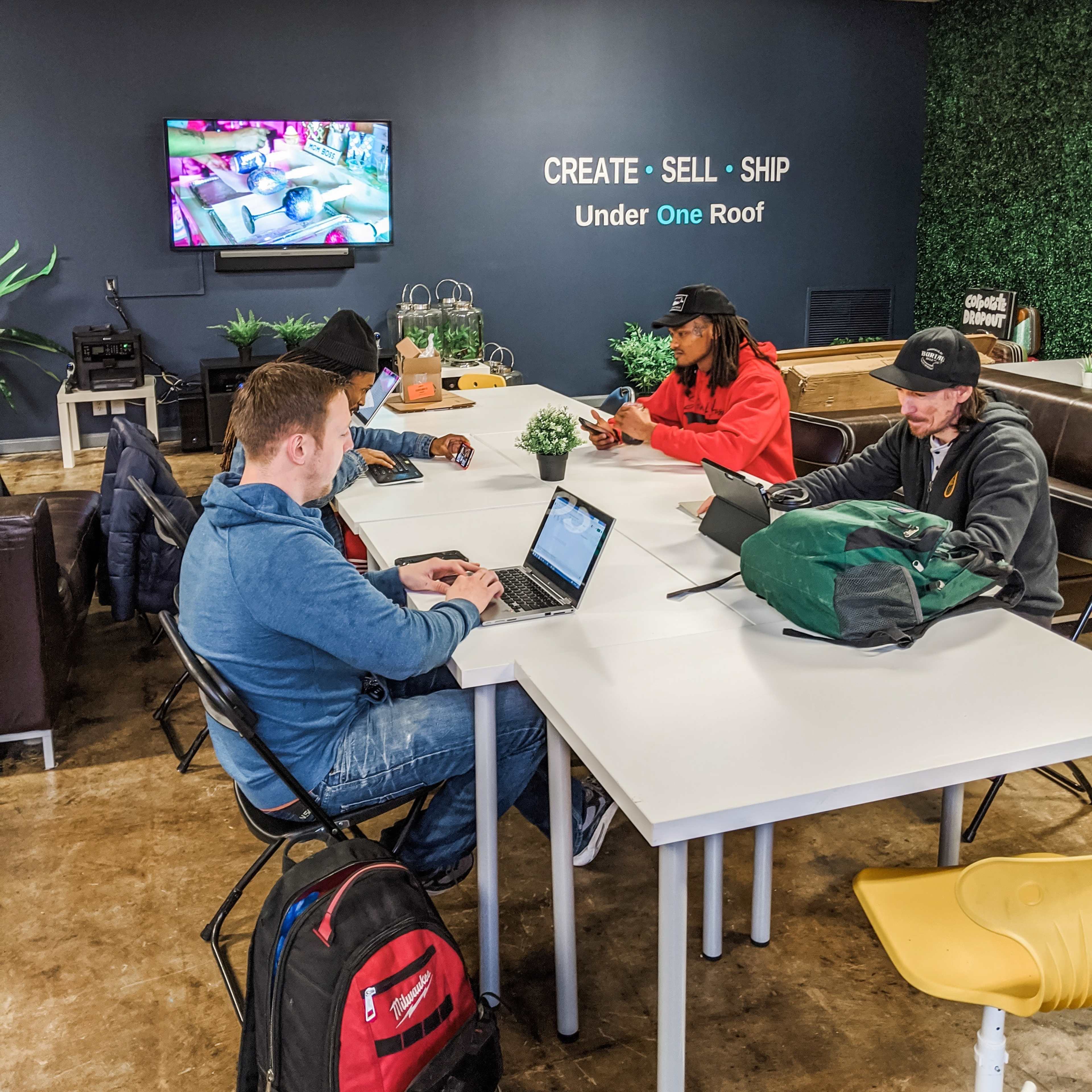 A group of five individuals is seated around a white table in a modern workspace, each engaged with their laptops and phones, while a television displays a colorful image on the wall.