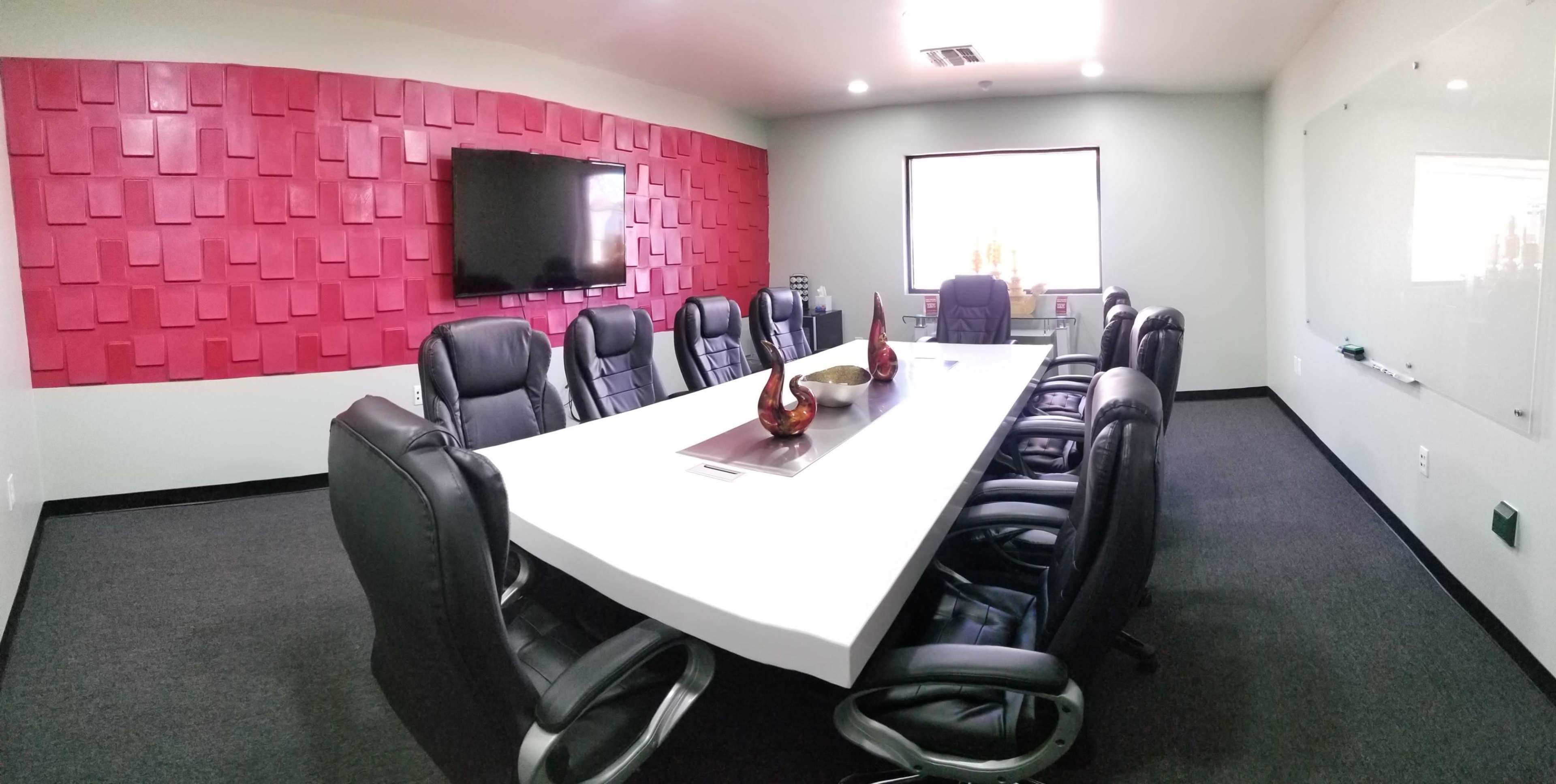 A modern conference room features a large white table surrounded by black leather chairs, a wall-mounted TV, and a red textured wall.