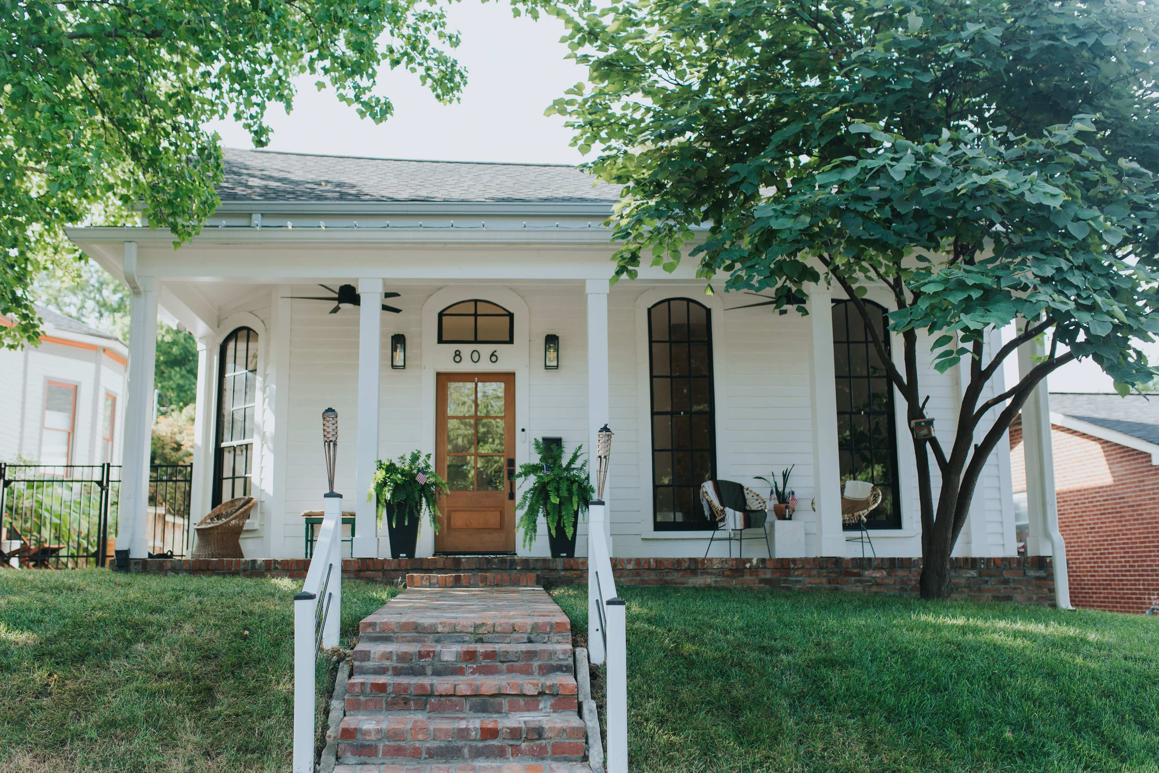 A white house with large windows, a wooden front door, and a brick pathway, is surrounded by greenery and features two chairs on the porch.