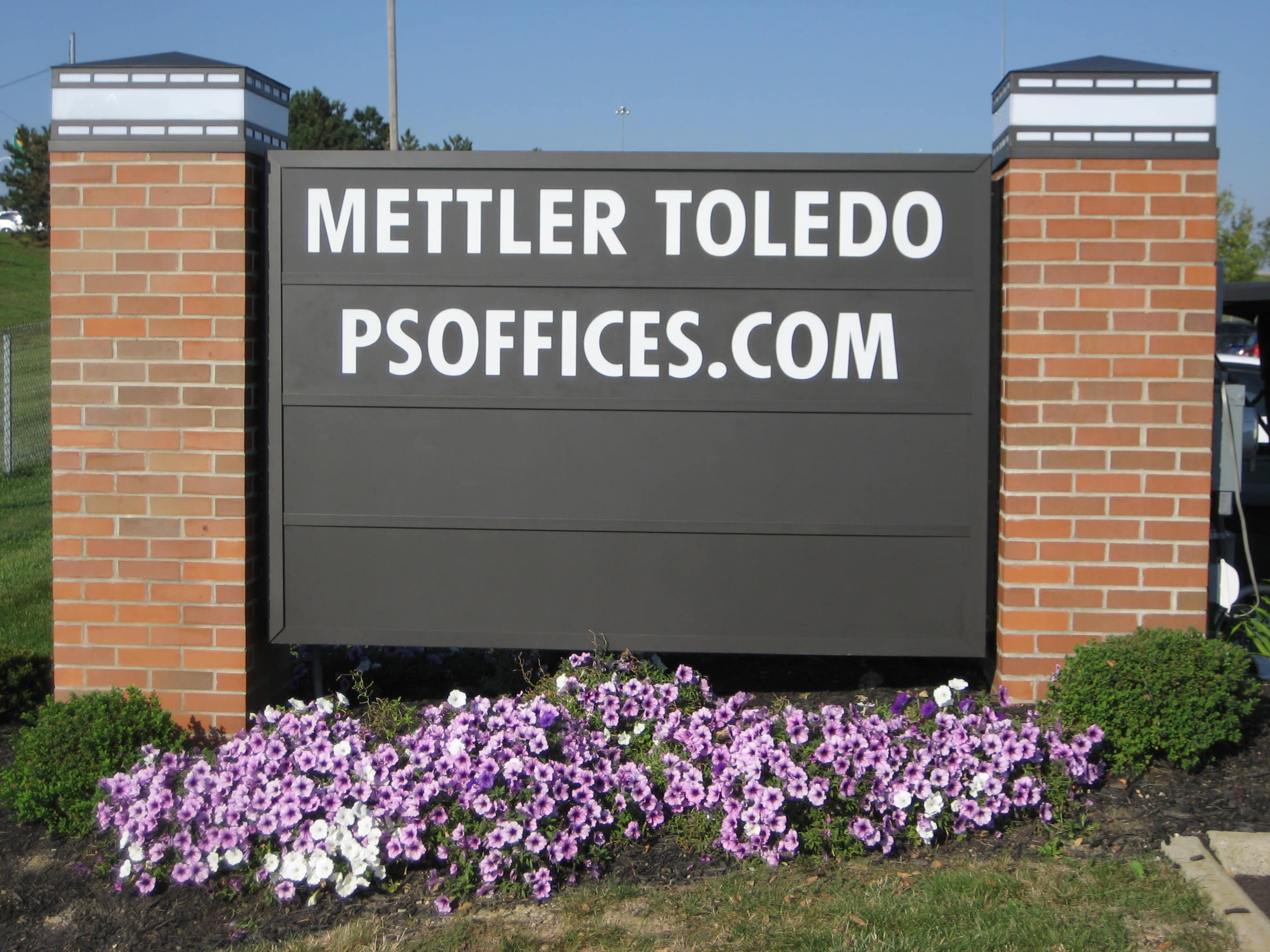 A sign for Mettler Toledo with the website psOffices.com is displayed in front of a flowerbed with purple petunias.