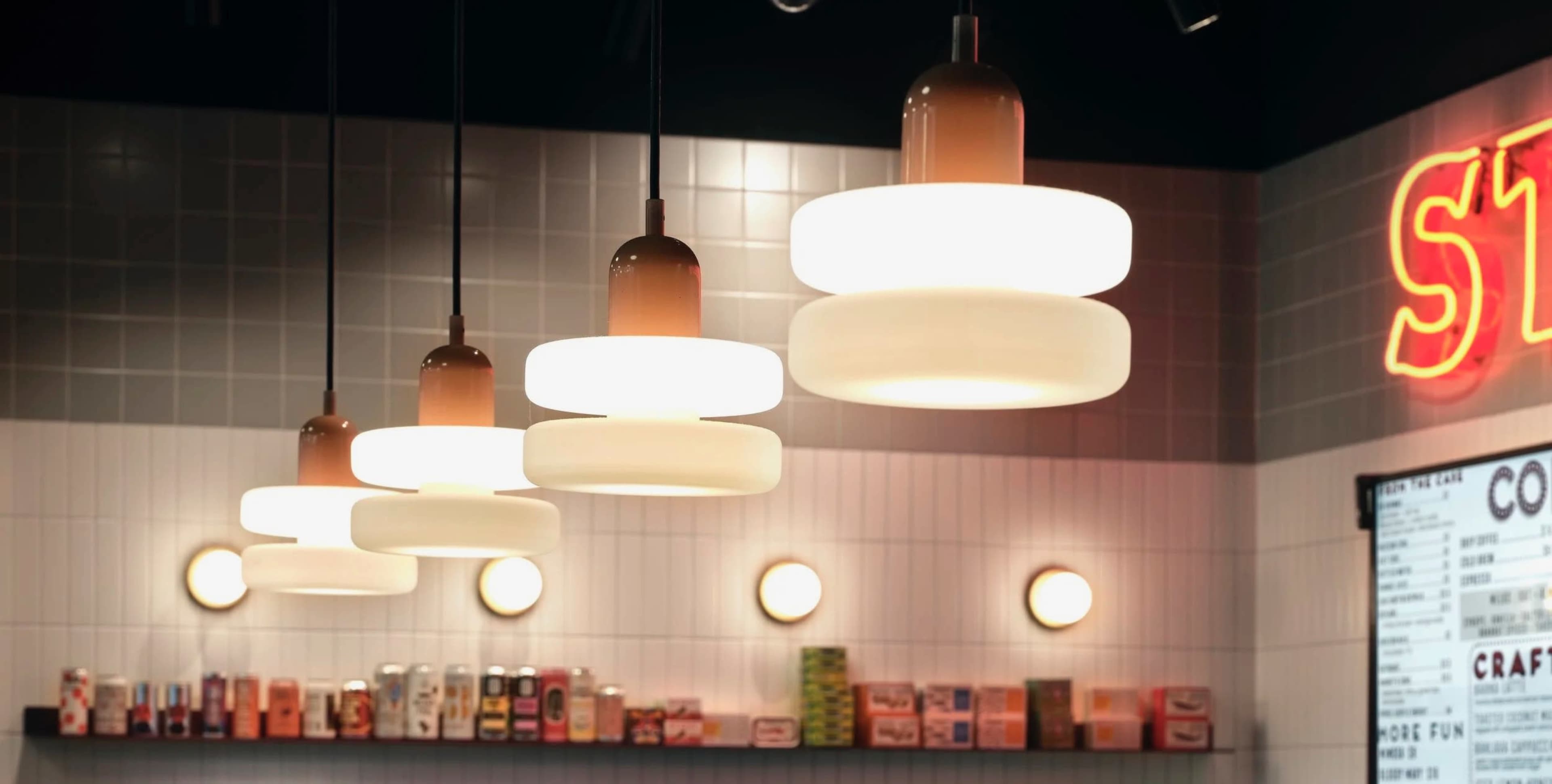 The image shows a modern café interior with pendant lights hanging from the ceiling and a wall adorned with a neon sign and shelves filled with various products.