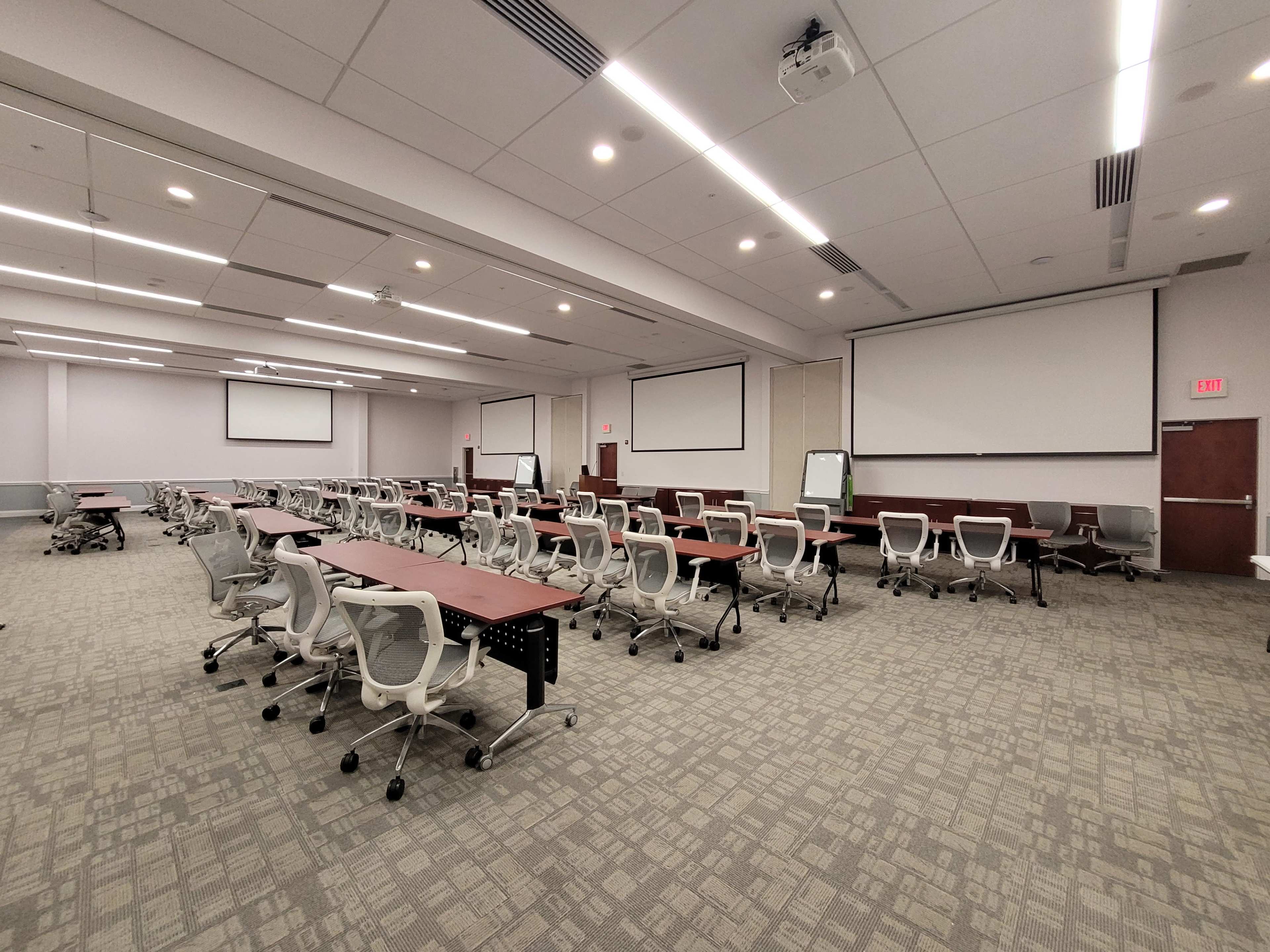 A modern conference room is set up with several rows of tables and chairs, equipped with projectors and large screens on the walls.