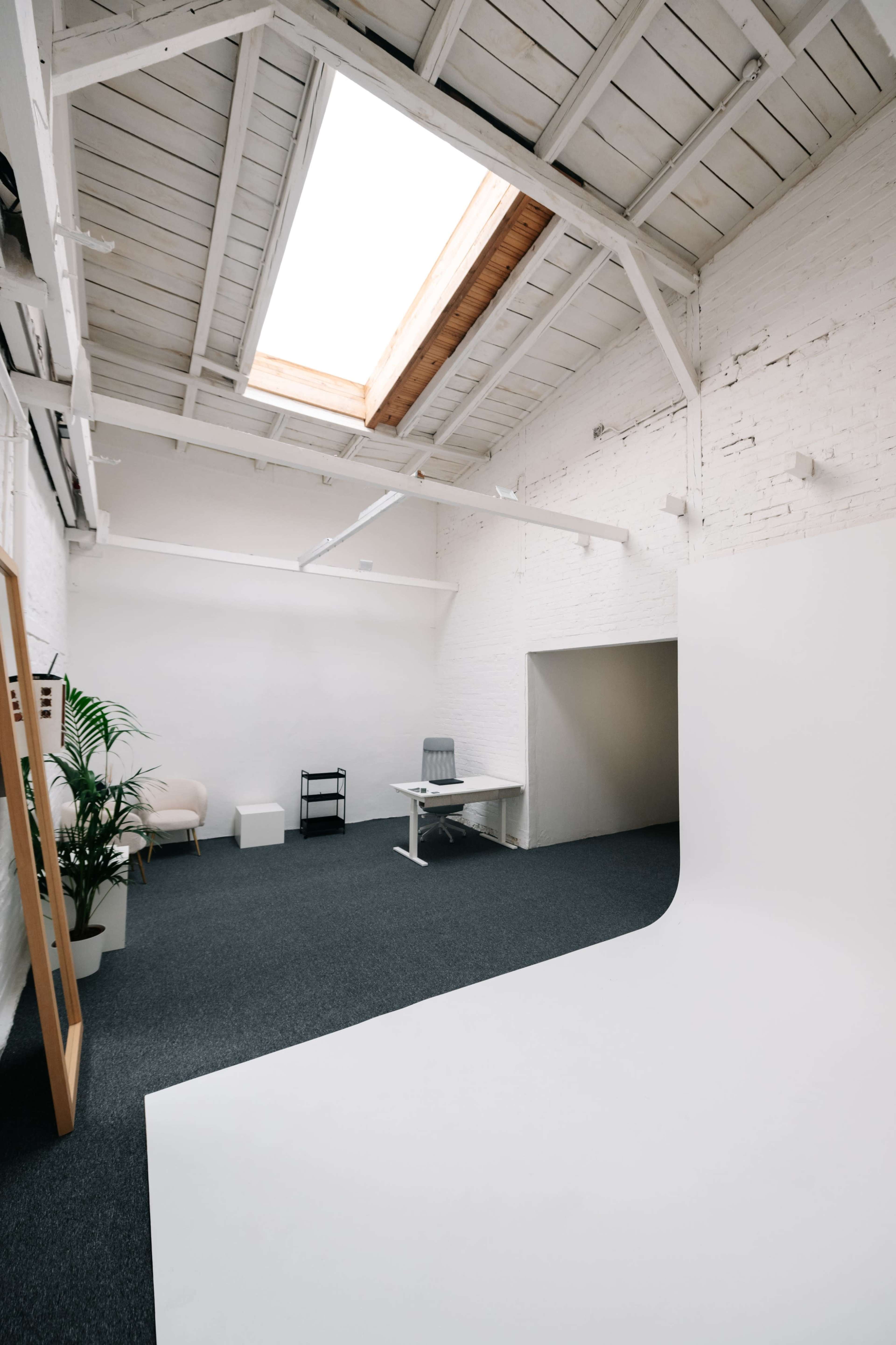 The image shows a minimalist studio space with white walls, a skylight, a desk, a chair, and a backdrop area.