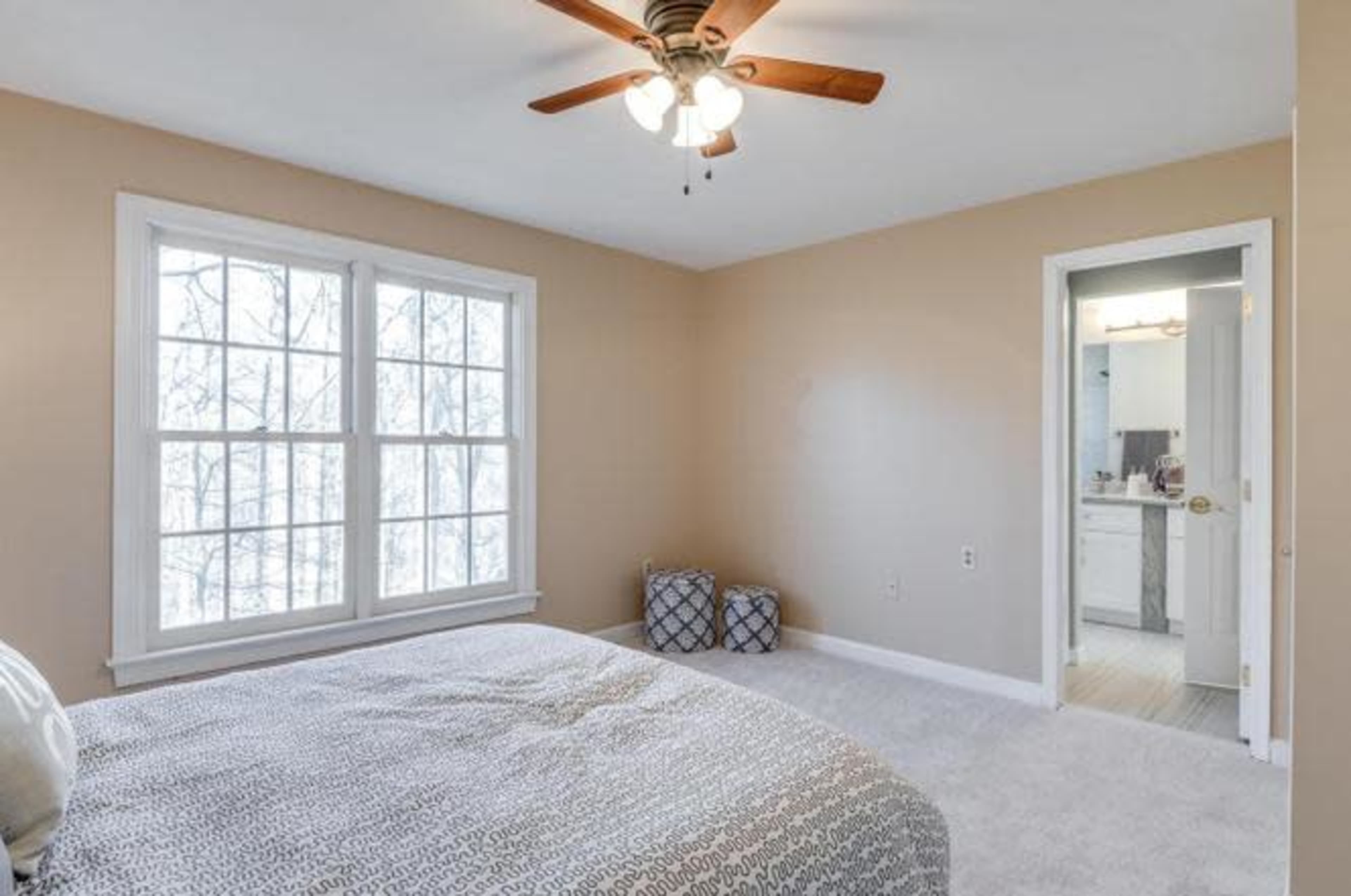 The image shows a simple bedroom with a bed, a ceiling fan, and a doorway leading to a bathroom, illuminated by natural light from a large window.