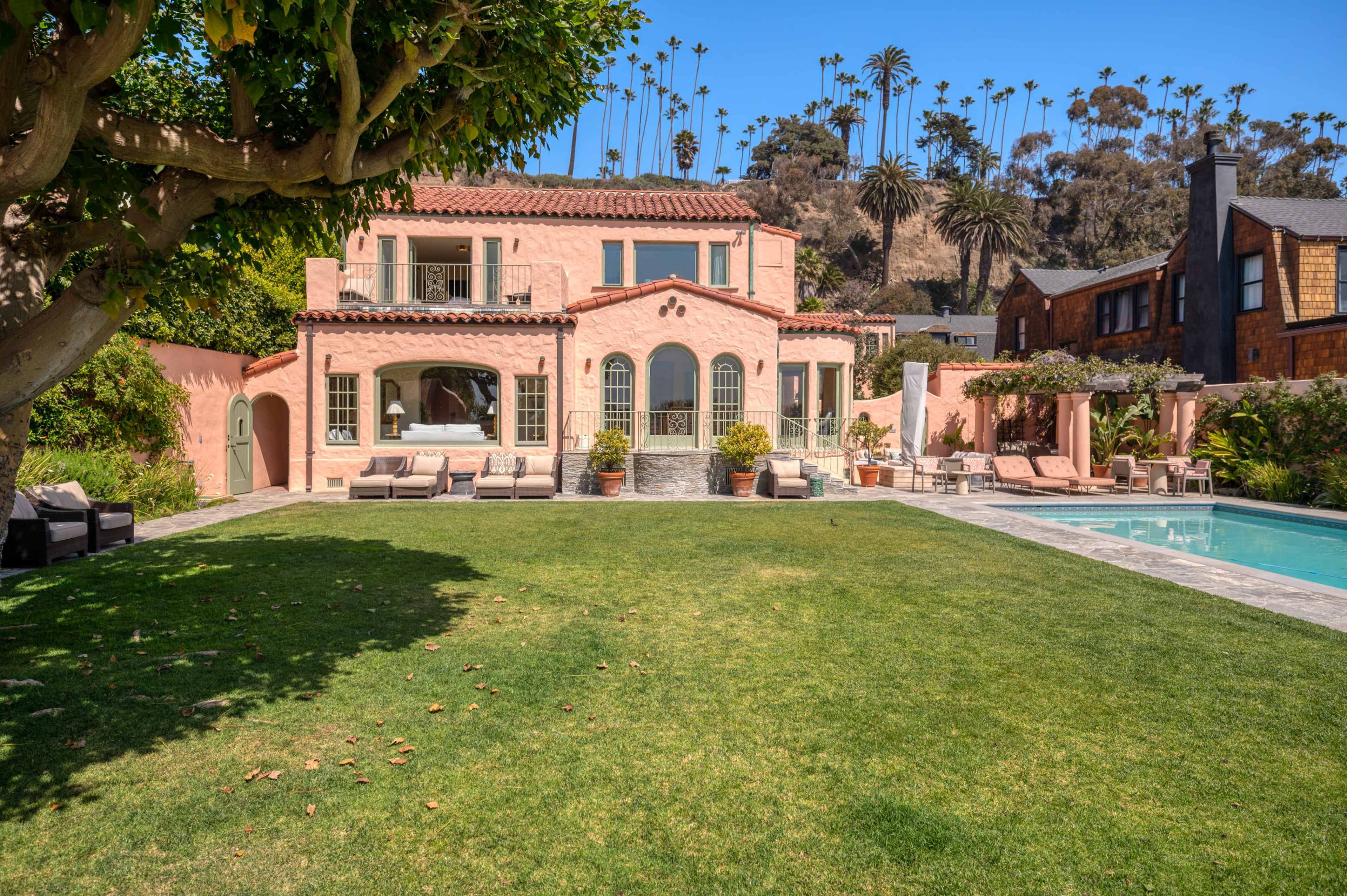 A pink two-story villa with arched windows and a swimming pool is surrounded by a landscaped yard and palm trees.
