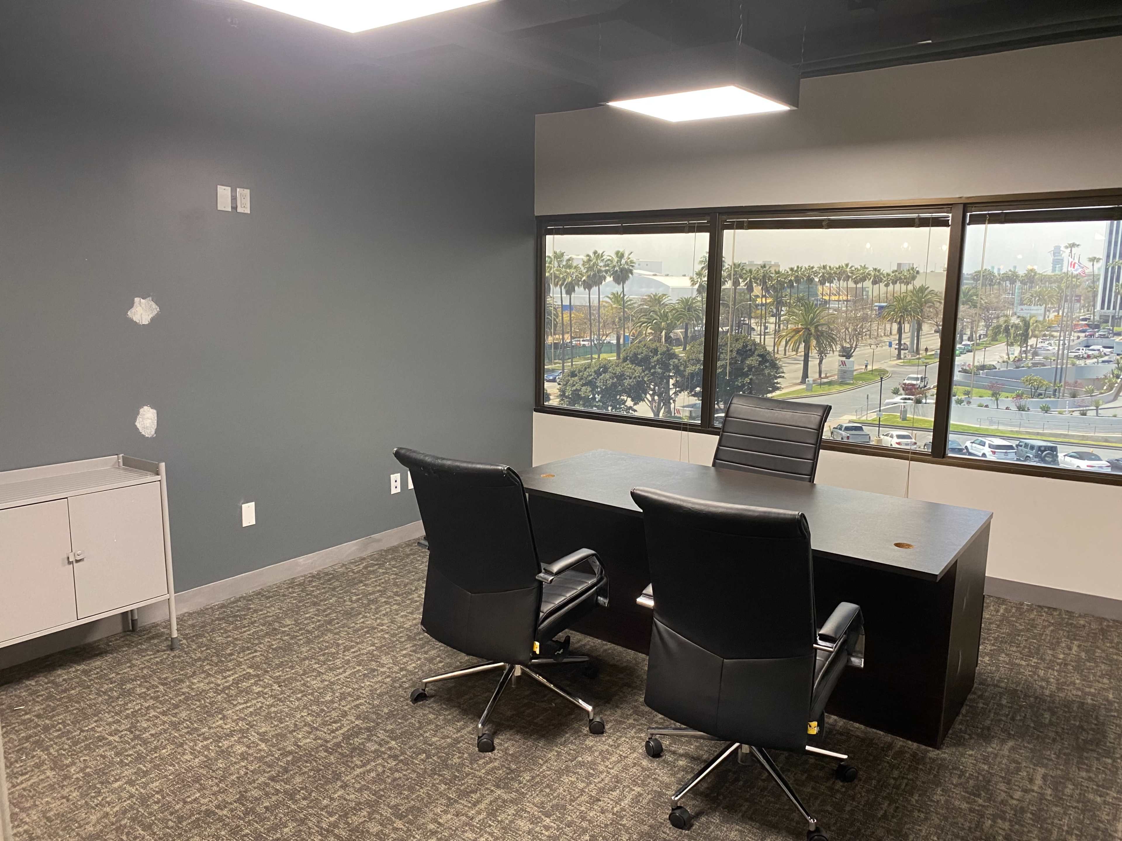 A conference room features a large table with two black office chairs, a gray cabinet, and windows overlooking a parking lot and palm trees outside.