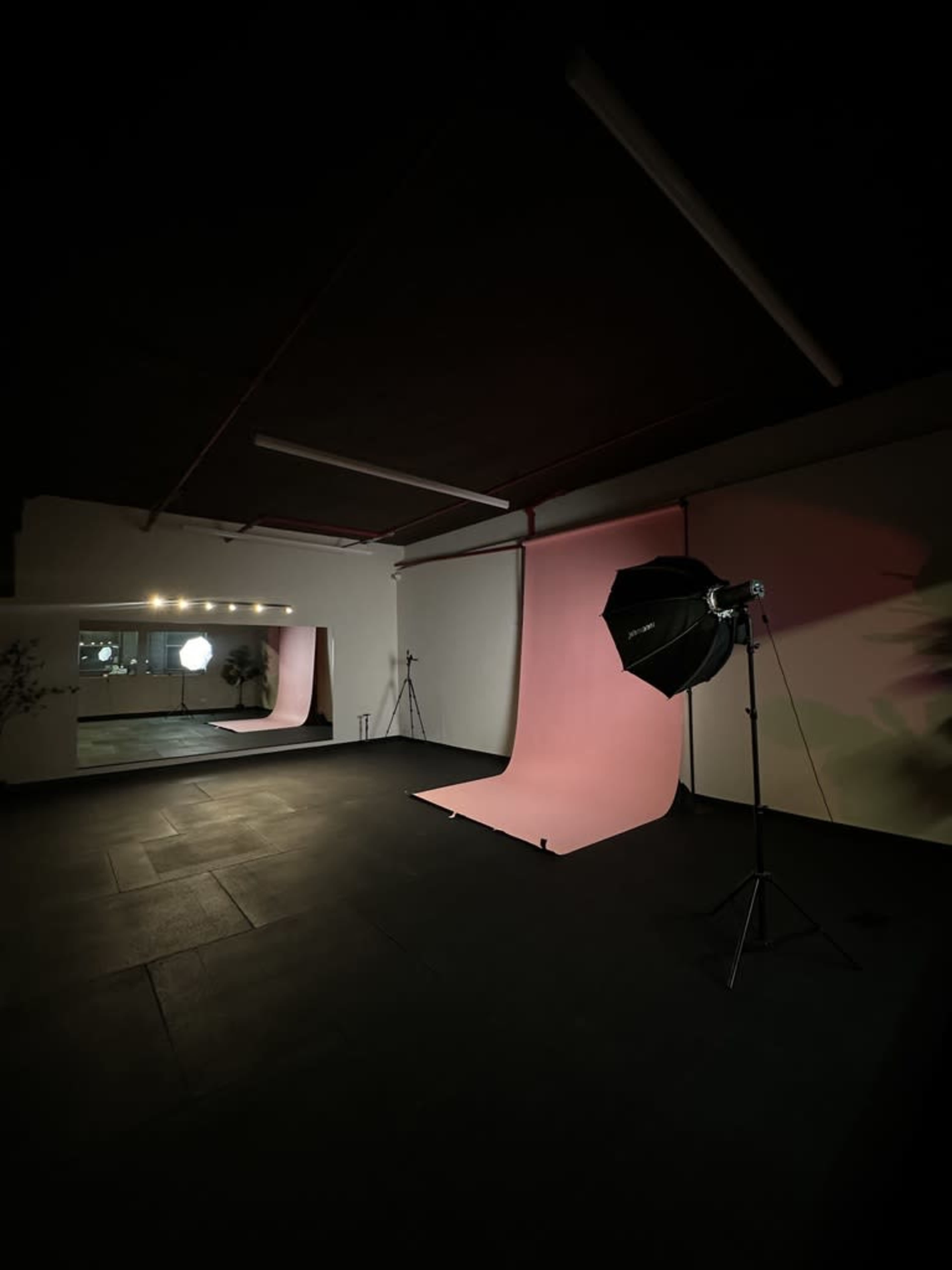 The image shows a dimly lit photography studio featuring a pink backdrop, a lighting setup on a stand, and a mirror reflecting light.