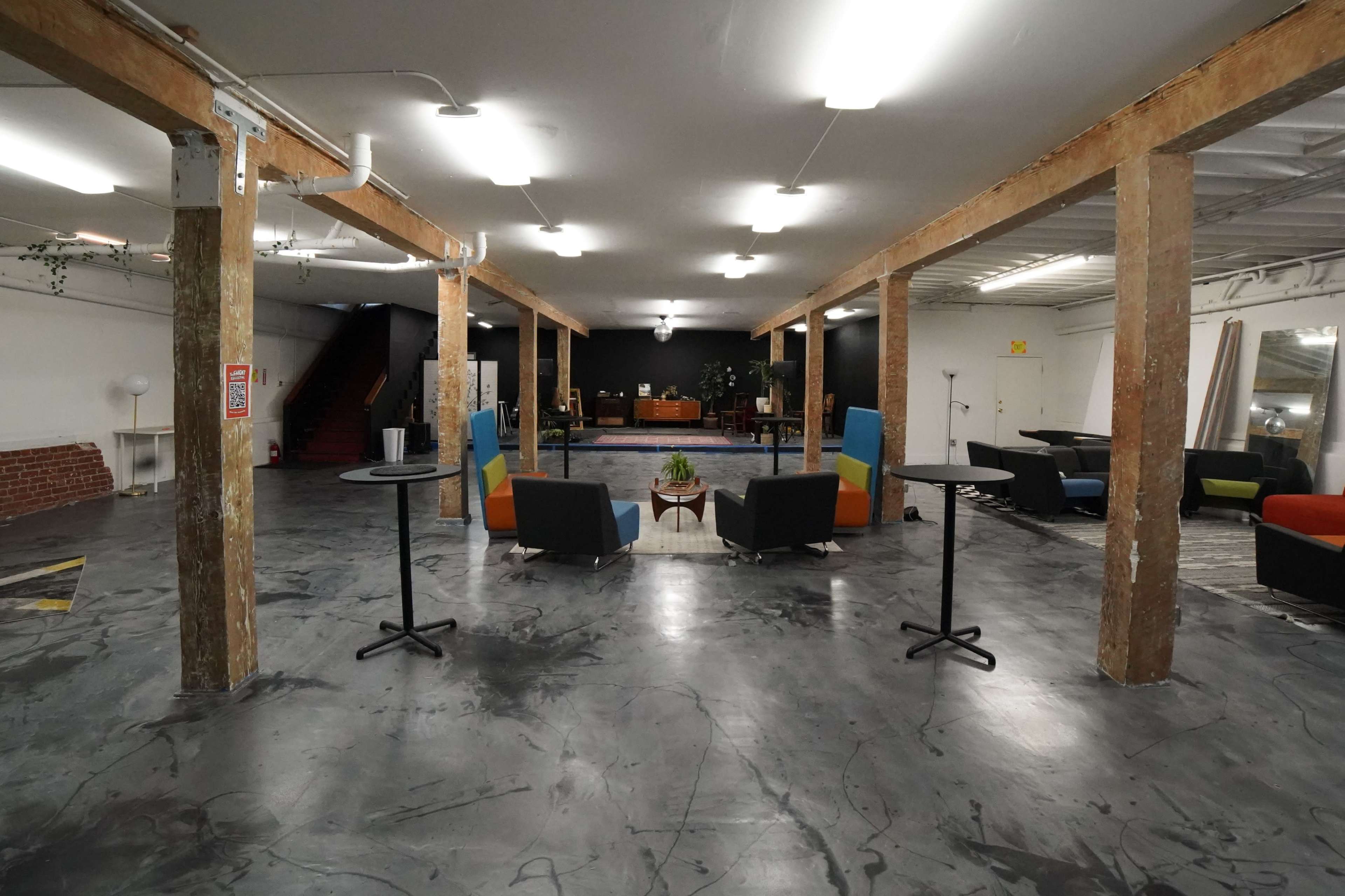 An industrial-style open space with wooden columns, concrete flooring, and various seating areas arranged around a central coffee table.