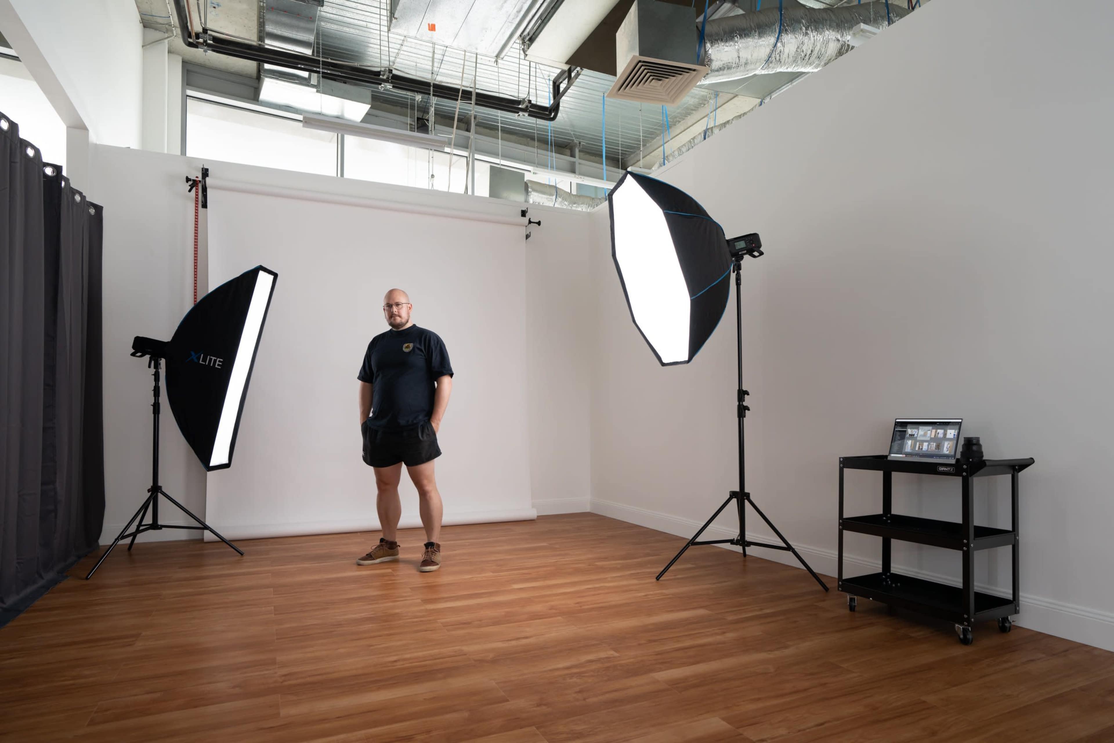 A man stands in a photography studio equipped with softbox lights and a portable cart with photographic equipment.