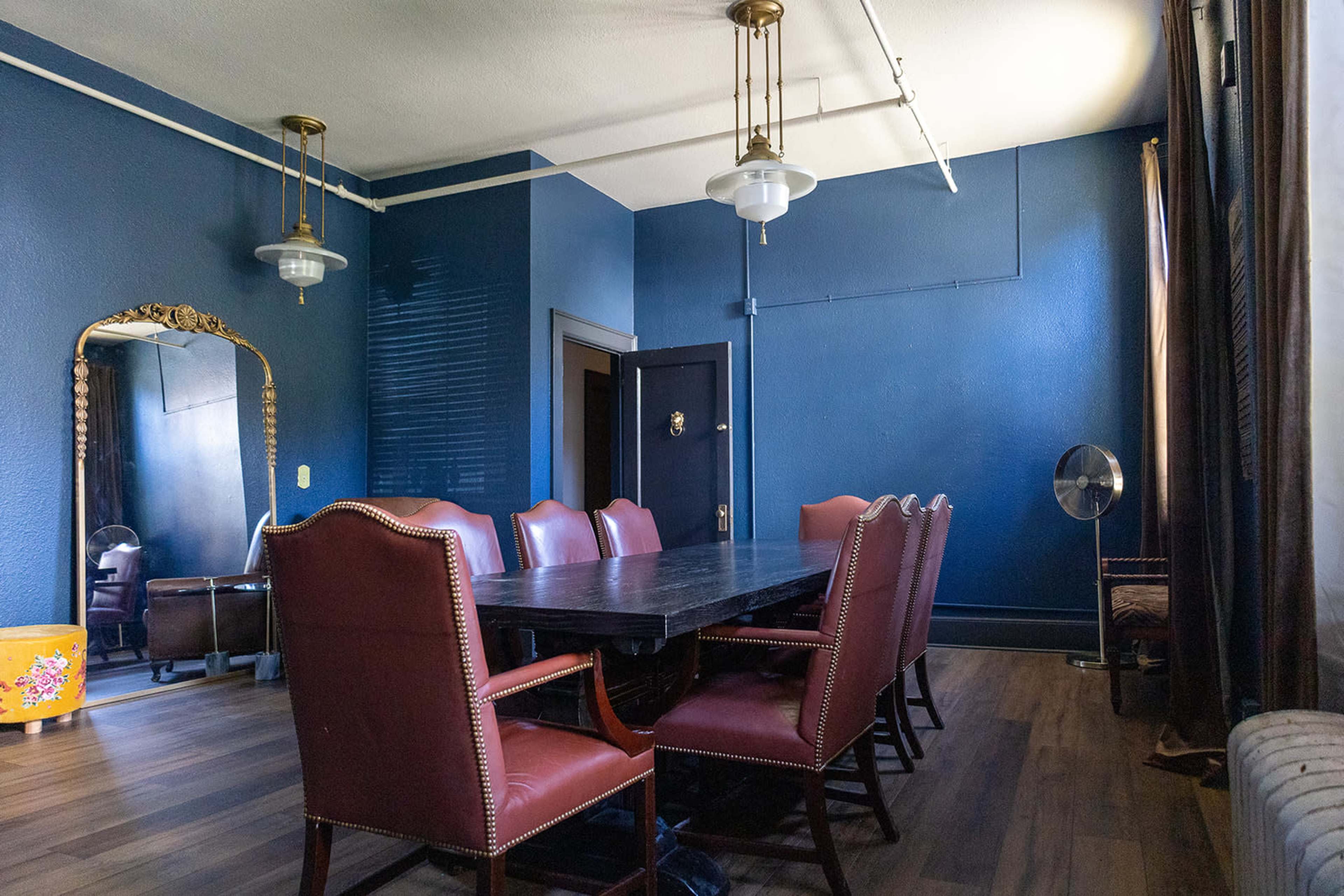 A dining room features a large dark wooden table surrounded by eight red leather chairs, with a mirror and fan against a deep blue wall.