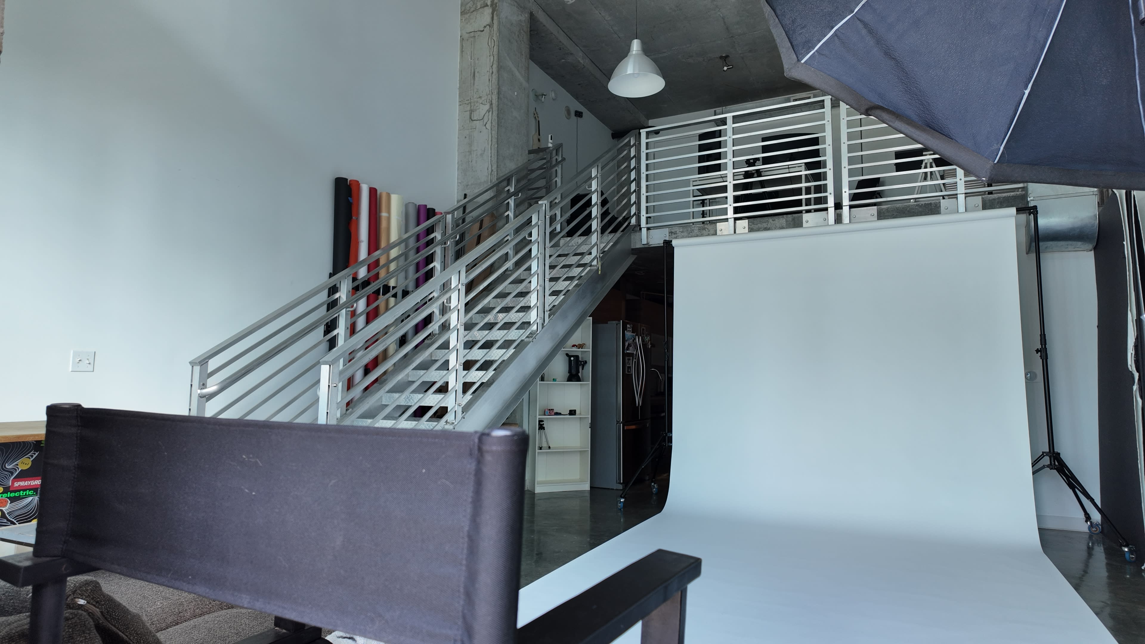 The image shows a modern studio space with a staircase leading to an upper level, a white backdrop set up for photography, and a director's chair positioned in the foreground.
