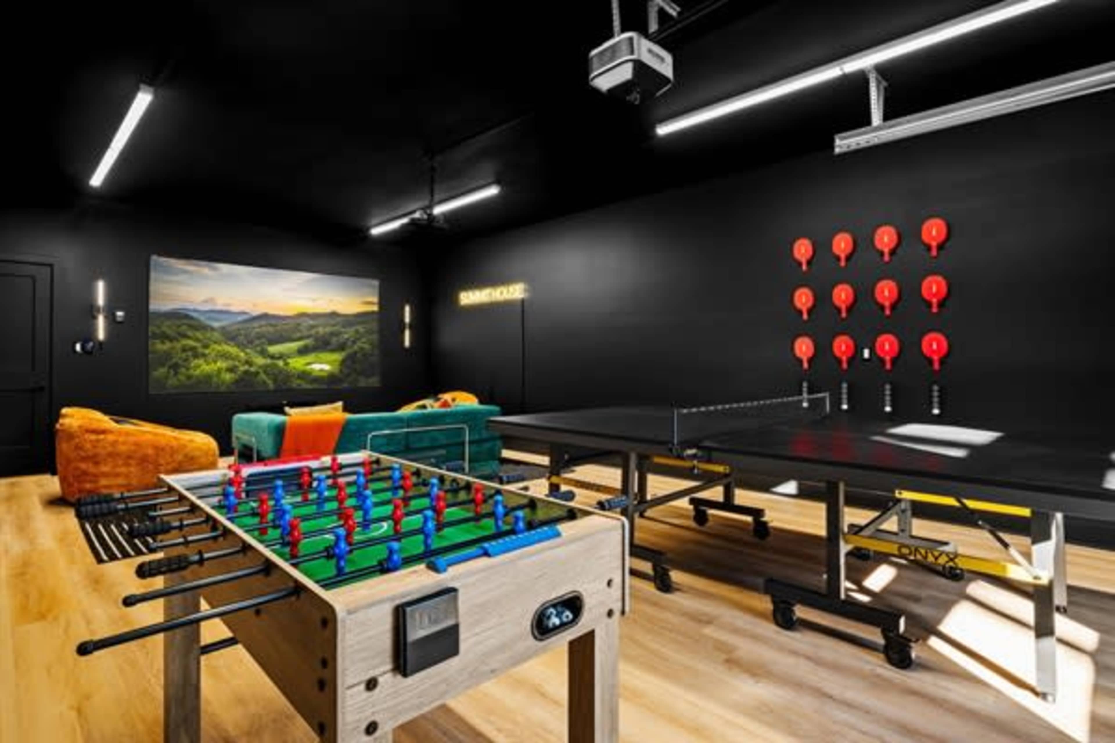 The image shows a recreational room featuring a foosball table, a ping pong table, bright seating, and a large wall-mounted screen displaying a scenic landscape.