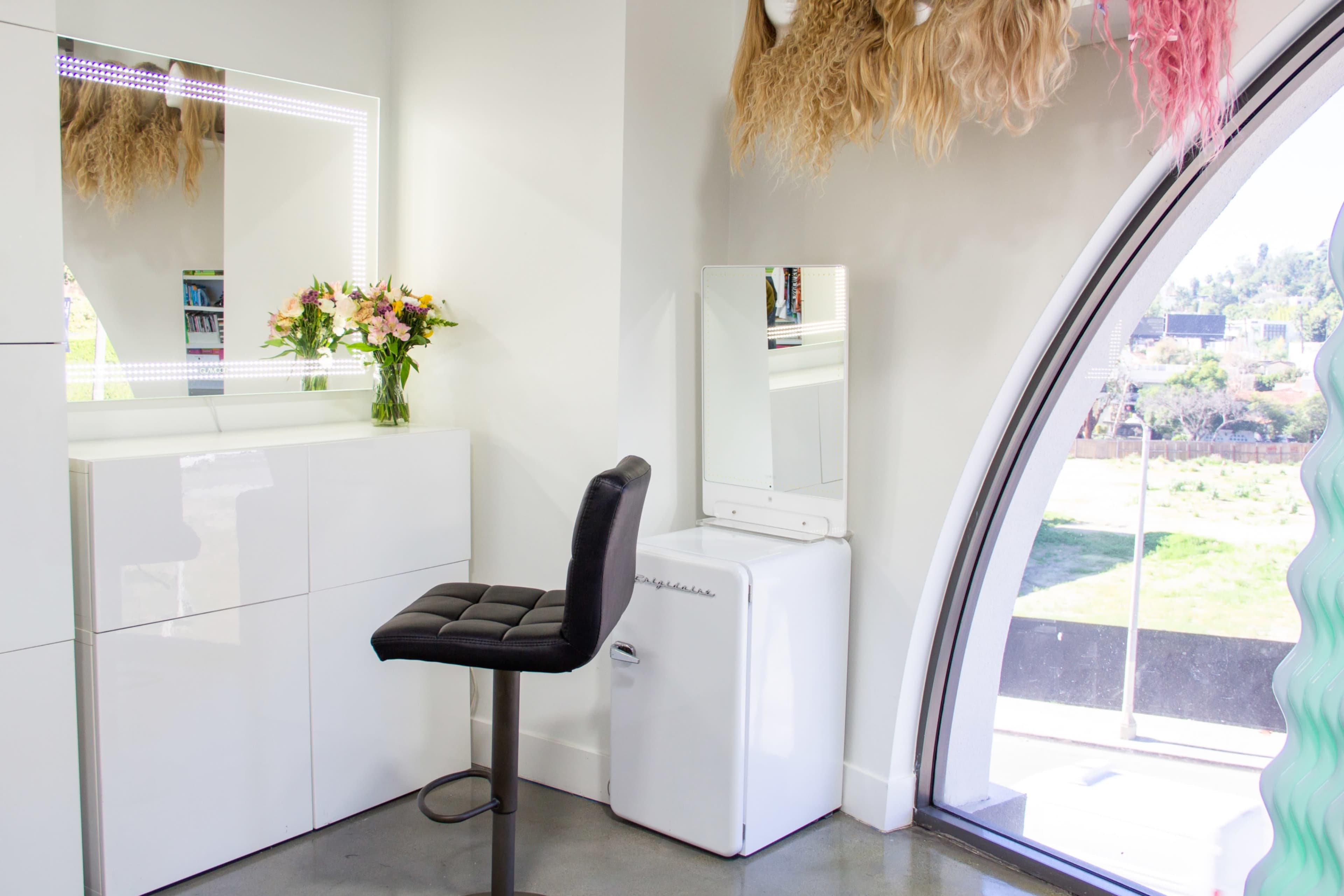 The image shows a modern grooming space featuring a mirror with LED lights, a small black stool, and a white refrigerator beside a large arched window with a view outside.