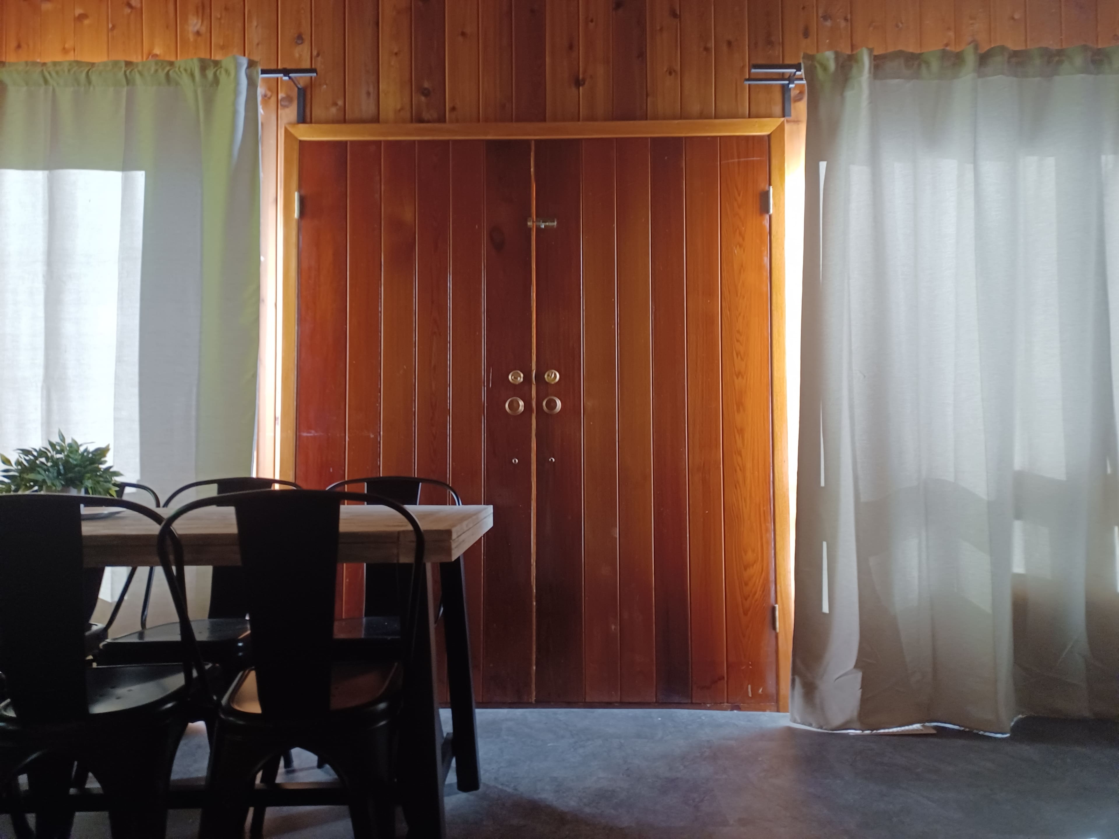A wooden double door is framed by light curtains, with a table and chairs positioned nearby in a room with wooden walls.