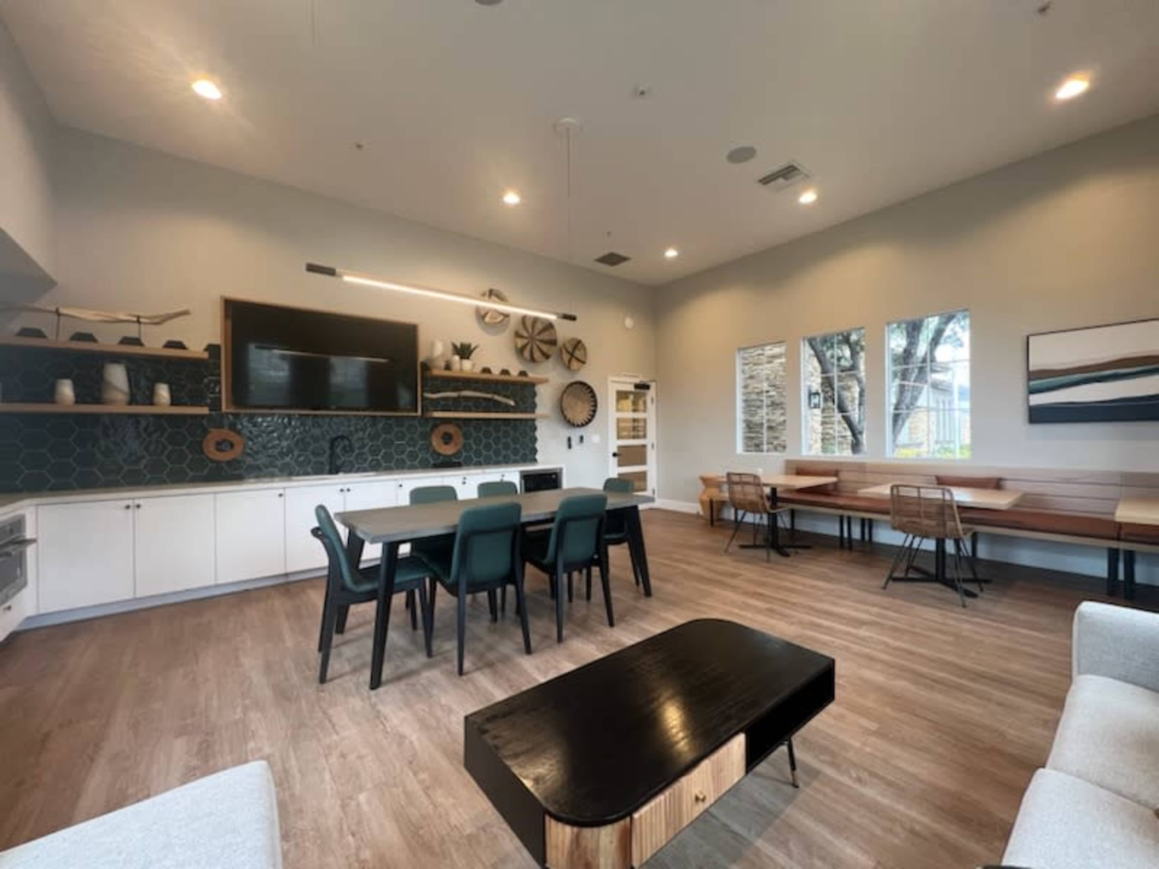 A modern common room features a dining table, seating area, and large windows, with a geometric patterned wall and various decorative items.