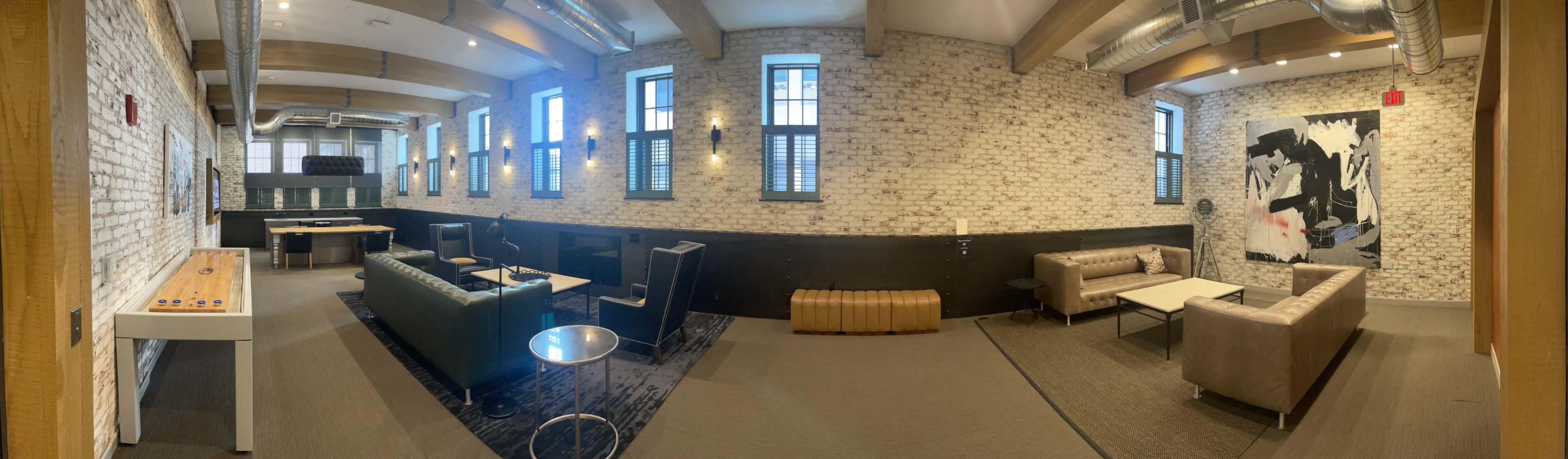 A spacious, modern interior with exposed brick walls, a lounge area featuring sofas and chairs, and a kitchenette in the back.
