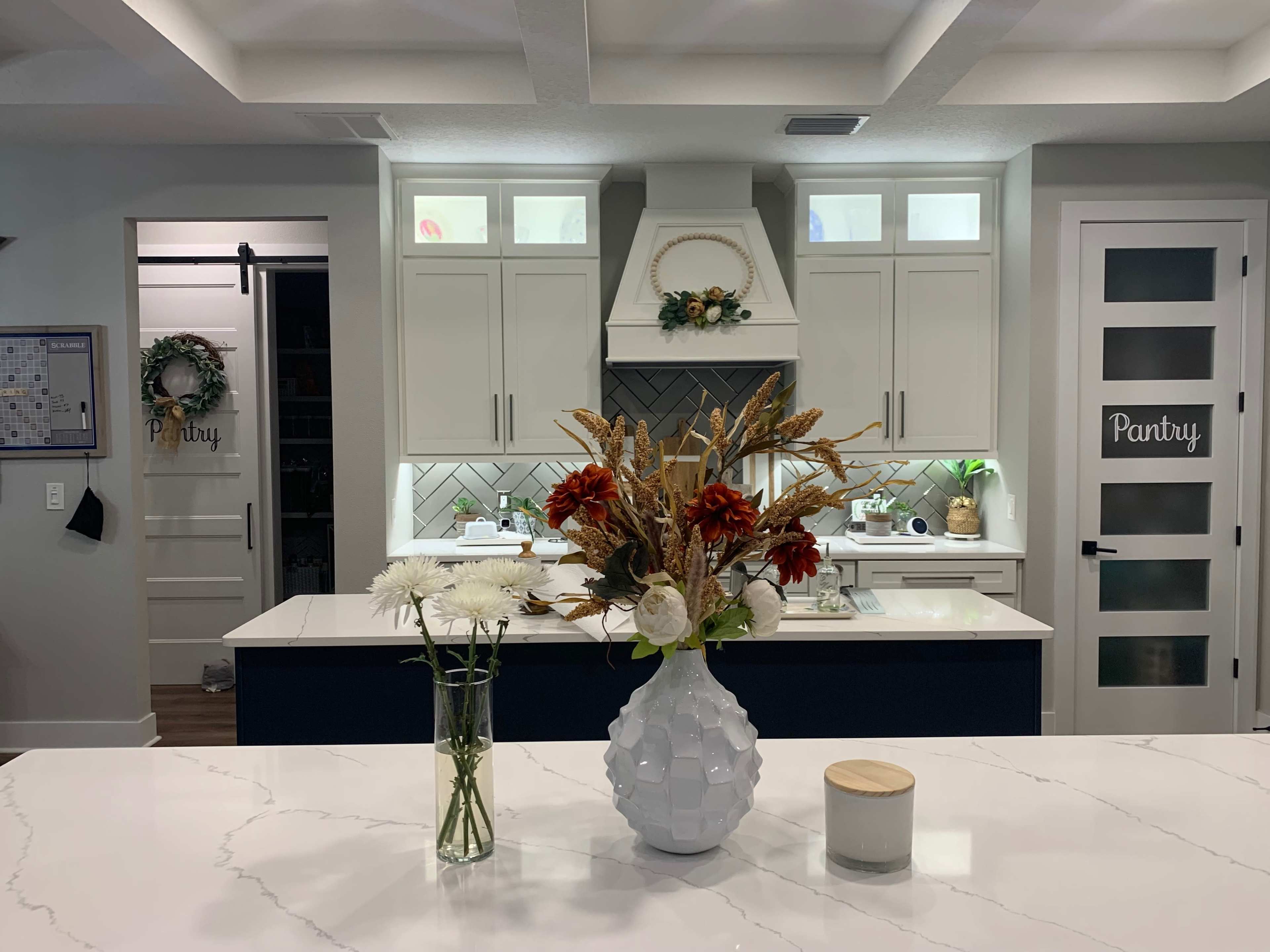 The image shows a modern kitchen with an island featuring a vase of flowers and a candle, while the pantry area is visible through a door on the right.