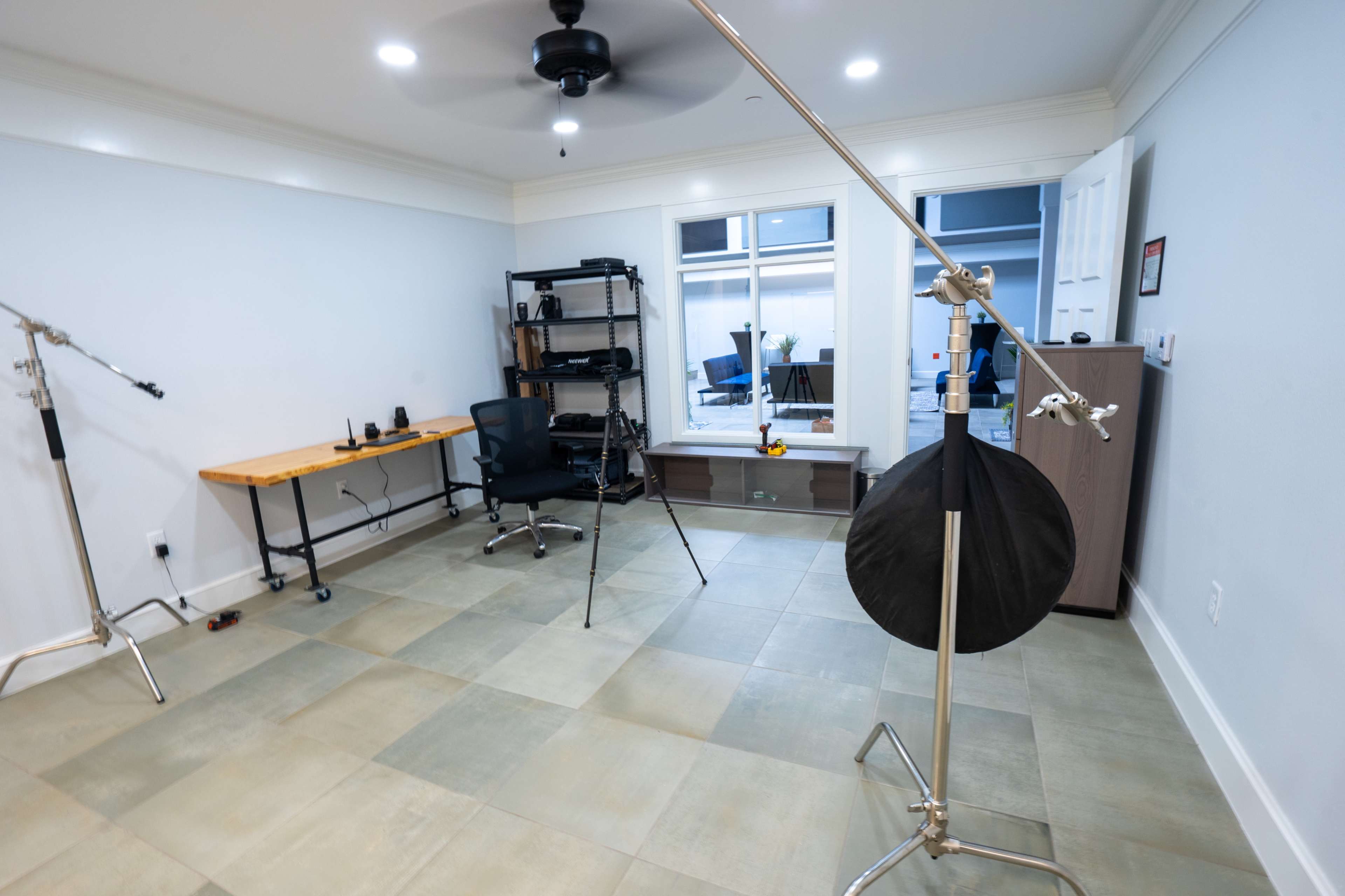 An empty room with light-colored tile flooring, featuring a wooden desk, black office chair, and several professional photography equipment stands.