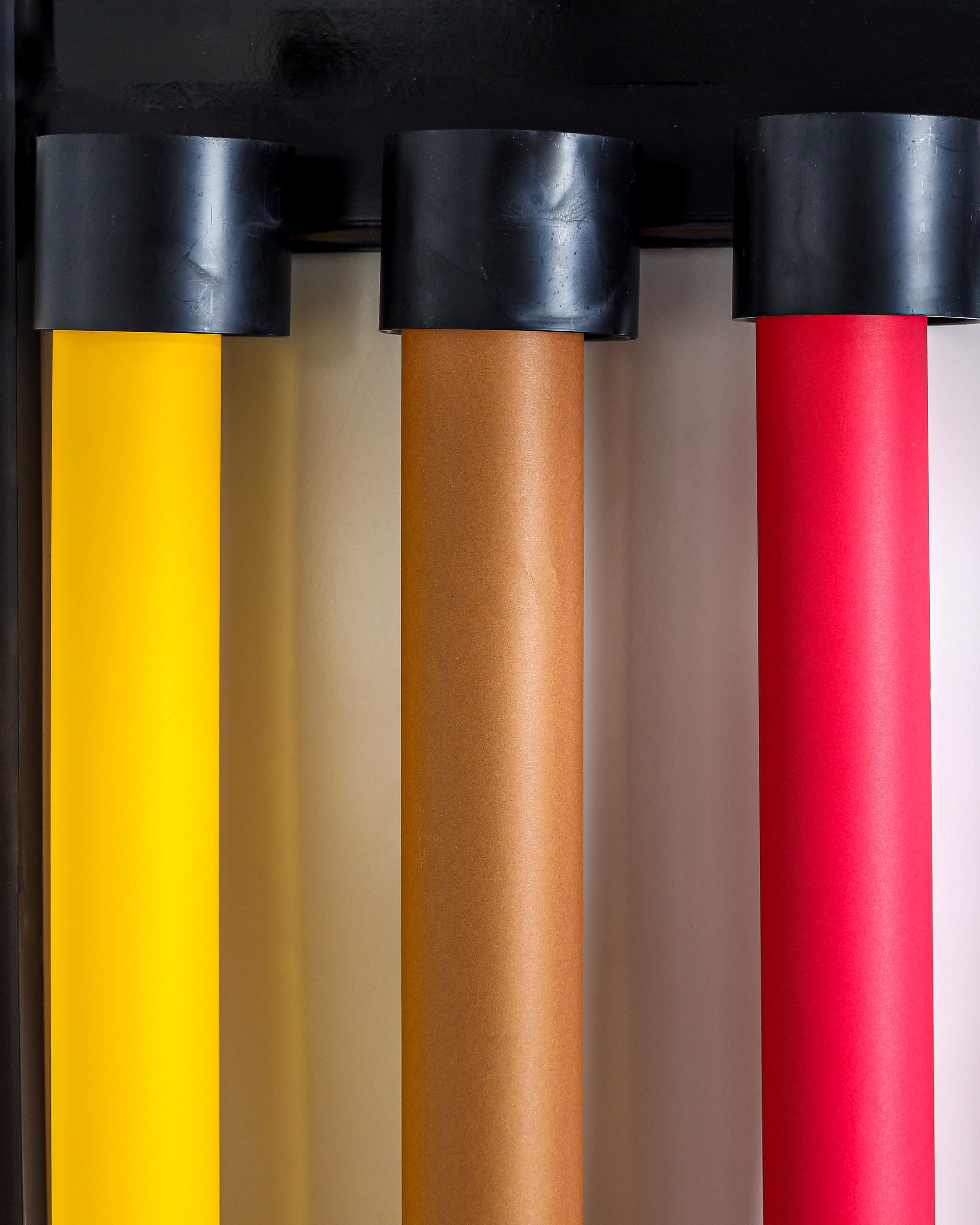 The image shows a row of five rolled paper sheets in various colors—yellow, brown, light beige, and red—resting against a black backdrop.