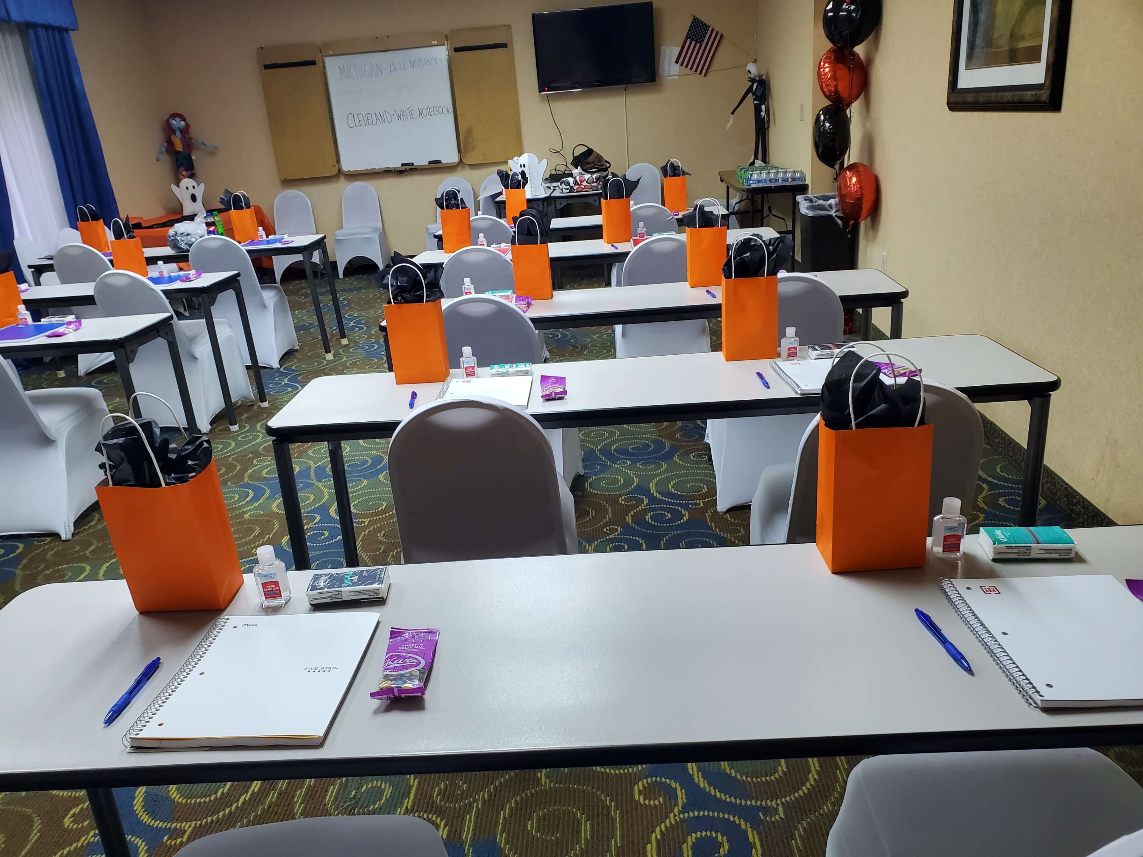 A classroom is set up with several tables, each adorned with orange gift bags, notebooks, pens, and hand sanitizer, under wall-mounted decorations and a television.