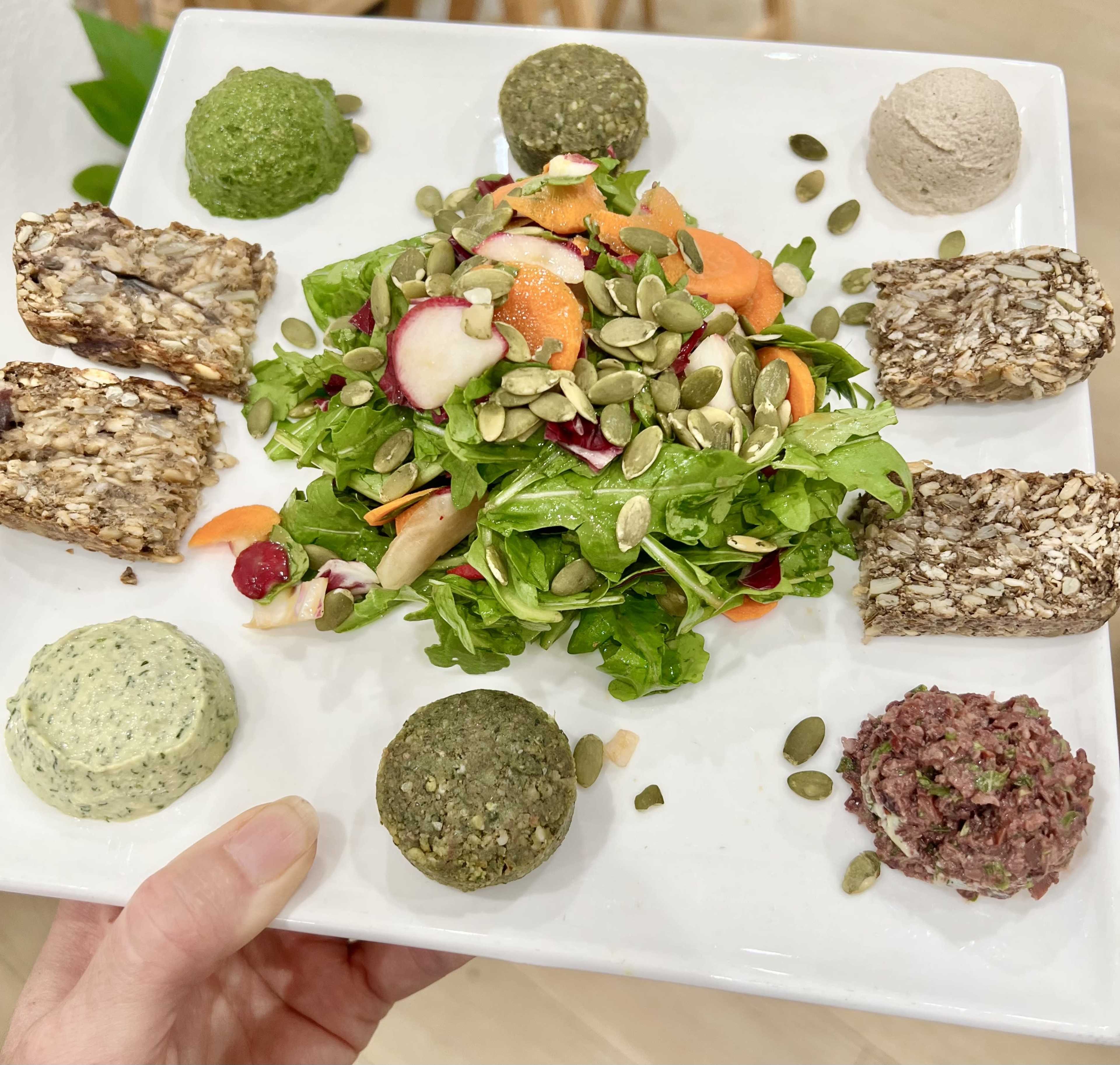 A person holds a square plate featuring a colorful salad with various toppings and six small circular portions of different spreads.